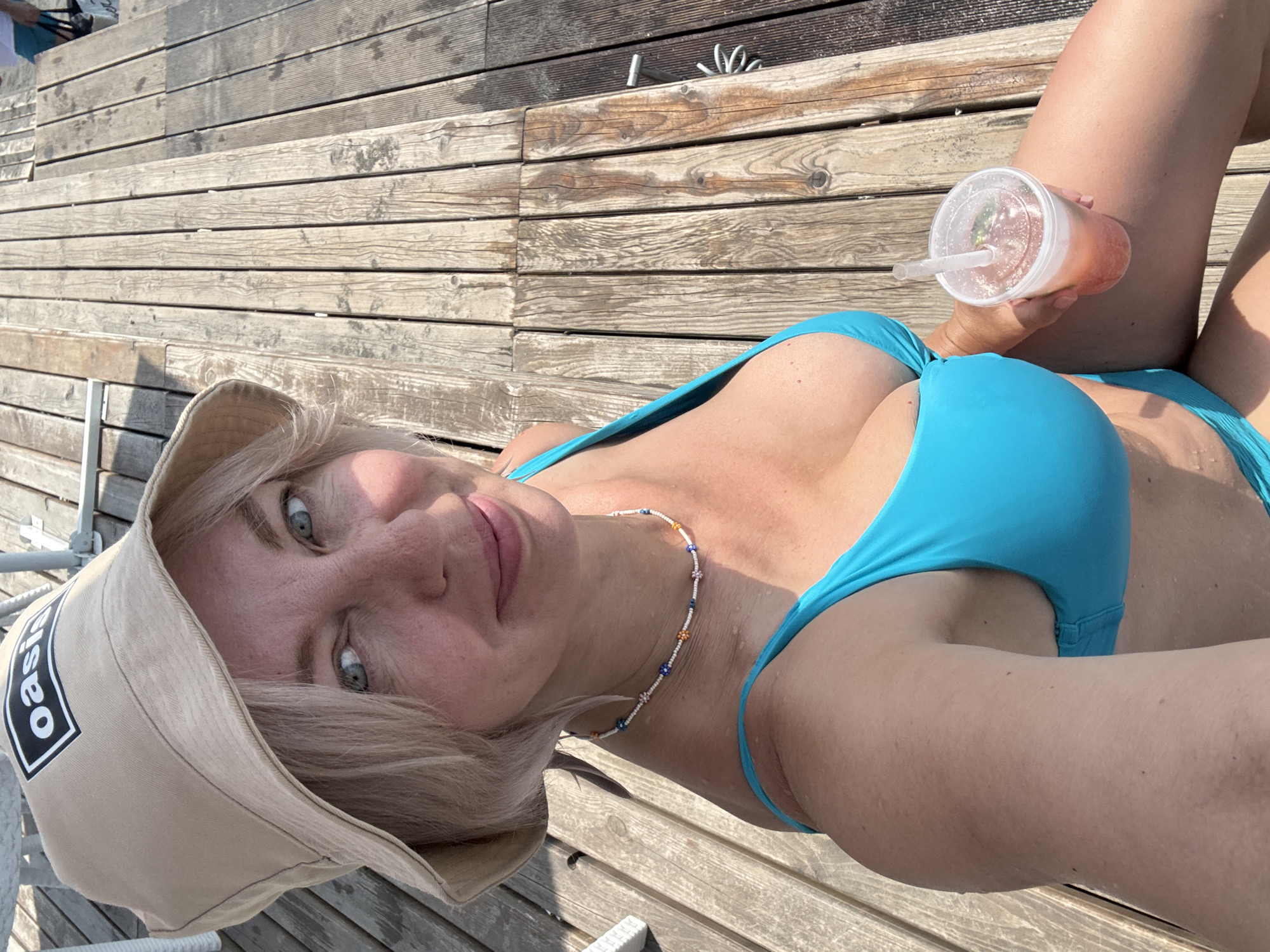A fair-skinned woman with blonde hair and blue eyes wears a tan bucket hat with an "Oasis" logo and a bright blue bikini. She is sitting on a wooden deck, holding a clear plastic cup with a pink drink and a straw. The deck appears to be outdoors, possibly near water, given the presence of a chair and the woman's attire. The lighting suggests it is daytime. The location is identified as Sirius, Russia.