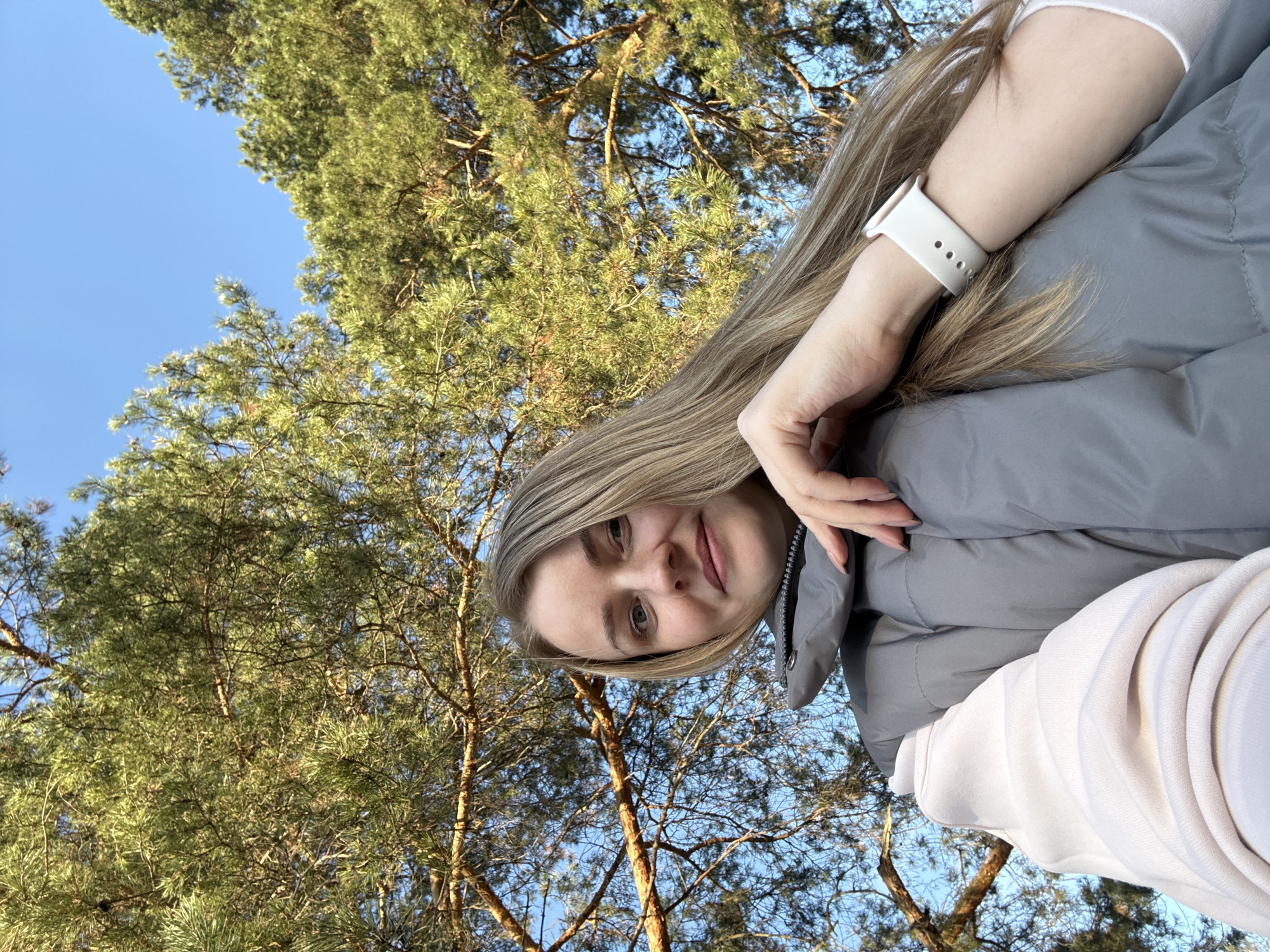 This media file is a low-angle selfie featuring a young woman looking towards the camera, set against a backdrop of a clear blue sky and a dense canopy of coniferous trees. The image is rotated, presenting the woman's face on the right side, looking slightly upwards.
The subject is a woman with long, straight, light blonde or brown hair, parted on the side. She has fair skin, light-colored eyes (likely blue or grey), and a natural lip color, with a soft, content expression. She is wearing a grey puffer jacket or coat over a light-colored (possibly white or light pink) long-sleeved top or hoodie, parts of which are visible. A white smartwatch with a silicone band is prominent on her left wrist. Her right hand is near her cheek, and her left arm is bent.
The setting is outdoors, likely in a forest or park. The background is dominated by tall pine or similar coniferous trees with vibrant green needles and visible brown branches and trunks. A clear, bright blue sky is visible between the tree branches, indicating sunny weather. The bright illumination on the tree tops suggests it is daytime, possibly mid-morning or afternoon.
The overall scene conveys a sense of tranquility and connection with nature. There is no visible text in the image. The location context provided is Chaikovsky, Russia, which aligns with the depiction of coniferous forests common in such regions.