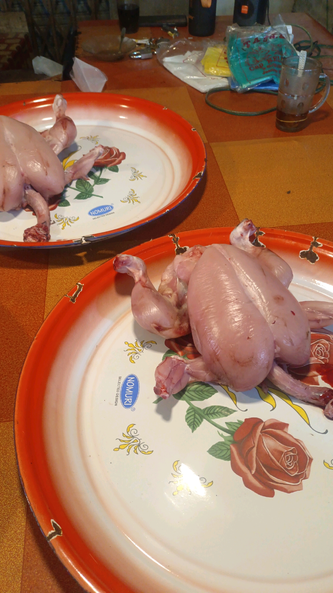 The image shows two raw chickens prepared for cooking, placed on separate decorative trays. The trays have a white base with orange rims and feature floral patterns, specifically roses, and a logo that reads "NOMURI SELECTED DESIGN". One chicken is positioned towards the foreground of the larger tray, with its legs spread out. The other chicken is on a tray in the background.
The background suggests a domestic setting, possibly a kitchen or dining area. There are various objects on the table, including plastic bags, a glass with a beverage and a spoon, and other unidentifiable items. The surface of the table appears to be a checkered pattern of orange and brown squares.
The image does not provide any direct visual cues to pinpoint its exact location within Padang Panjang, Indonesia. However, the presence of raw chicken prepared in this manner is common in Indonesian households, particularly for culinary traditions. The overall scene depicts a preparatory stage for a meal.