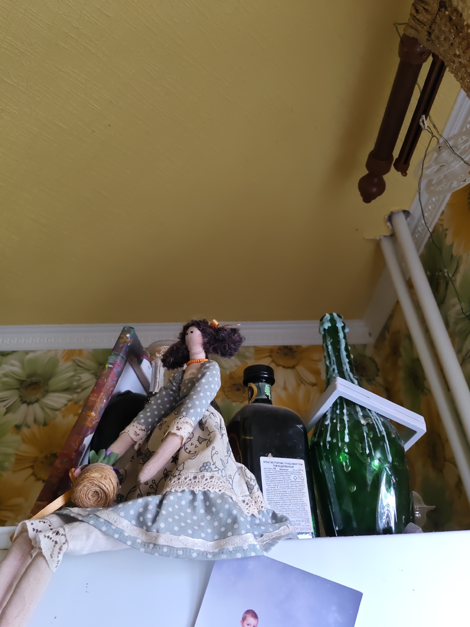 The image depicts a shelf with several objects, including a doll and two bottles. The doll is handmade, dressed in a blue and white polka dot dress with lace trim. It is positioned on the left side of the shelf. To the right of the doll are two bottles: a dark bottle with a label and a tall green bottle with white wax dripping down its sides. The background features floral wallpaper and a yellow ceiling. The scene appears to be indoors, likely in a domestic setting. There are no visible indications of the location being specifically in Rostov-on-Don, Russia.
