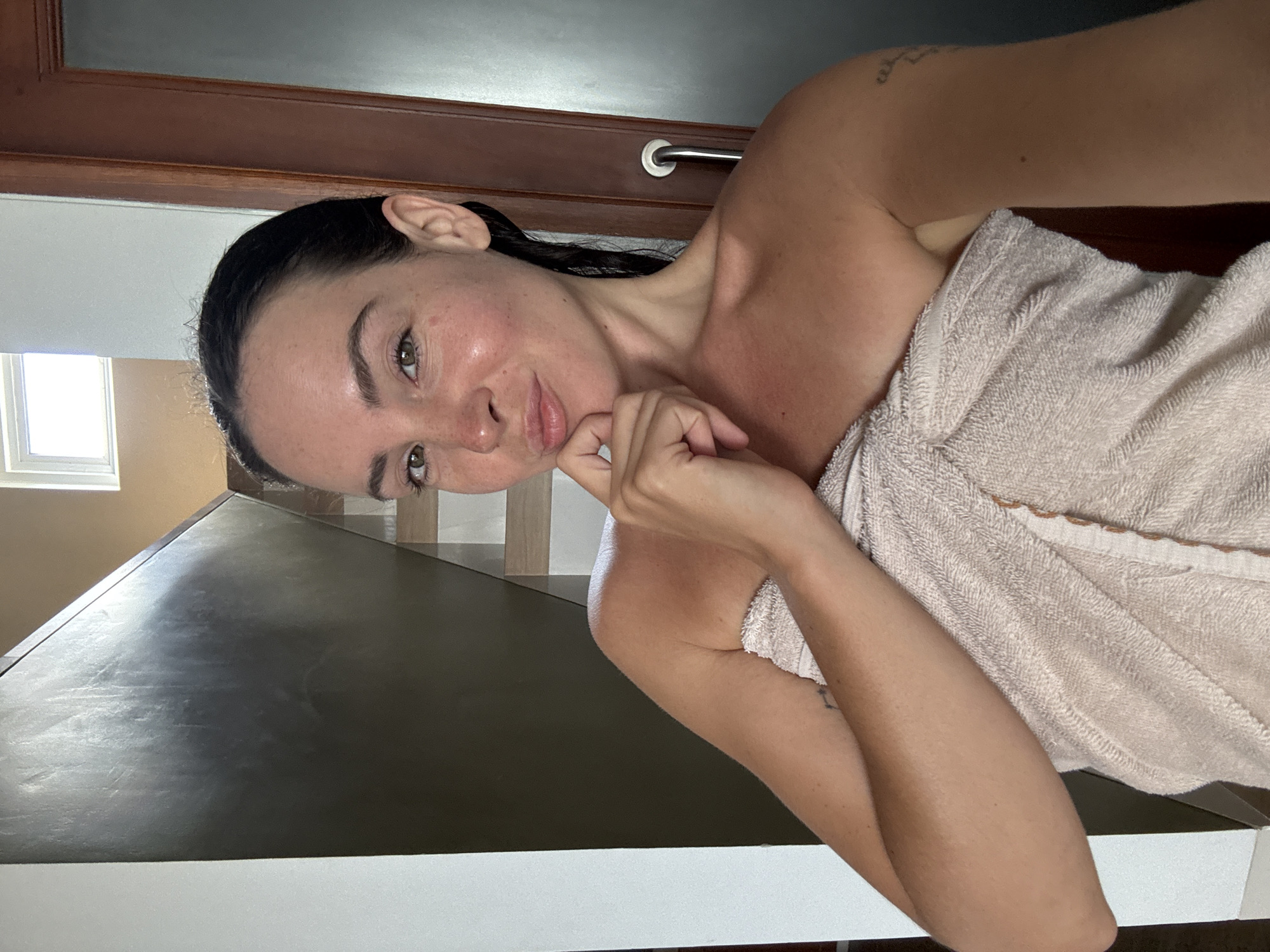 This is a portrait-oriented, low-angle, selfie-style photo of a woman. She has dark hair that is wet and pulled back from her face. She is wearing a light-colored towel wrapped around her body. Her right hand is resting on her chin, and she is looking directly at the camera with a slight smile. The background appears to be indoors, possibly a bathroom or spa area, with a wooden door and a towel rack visible. There is also a window with natural light coming through, and a dark, reflective surface in the foreground. The lighting suggests it could be daytime. The overall mood is relaxed and candid. There is no visible text in the image.