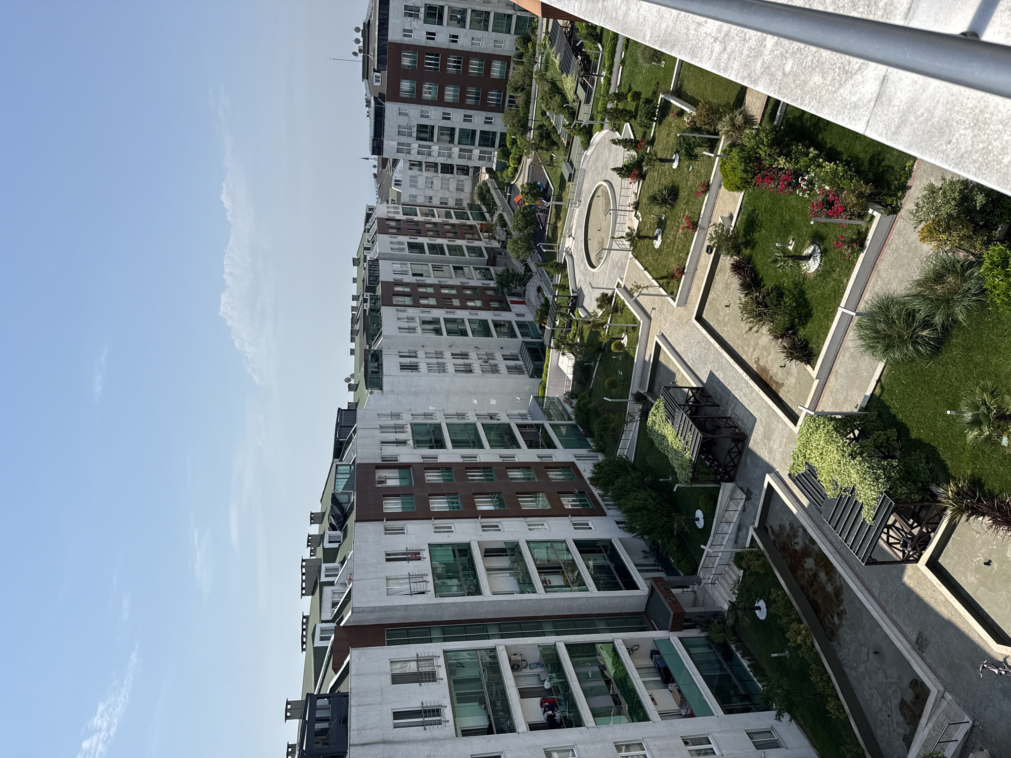 This media file presents a high-angle view of a modern residential complex in Beylikdüzü, Turkey, under a clear, bright blue sky with a few wispy clouds. The image captures several multi-story apartment buildings with contemporary architecture, featuring predominantly grey and white facades accented with dark brown panels and large glass windows and balconies. Many windows have security grates, and air conditioning units and satellite dishes are visible on the building exteriors and rooftops.
Between the buildings lies an expansive, meticulously landscaped communal garden area. This green space incorporates neatly trimmed lawns, a variety of small trees, bushes, and flowering plants, including vibrant red and white blooms. Paved pathways with geometric designs meander through the garden, connecting different sections. Notable features of the courtyard include a circular decorative fountain or water basin at the center of a paved area, and several long, rectangular water features or ponds with dark water. Wooden pergolas or trellises draped with greenery provide shaded areas, and benches are placed along the paths. In the background, a colorful children's playground with swings and a slide is visible.
The scene appears tranquil and devoid of any specific ongoing activity or event, though a bicycle is partially visible on a paved path. No people are clearly discernible in the image. The sunlight casts distinct shadows, indicating a bright daytime setting, likely morning or afternoon. No visible text is present in the image. The overall impression is one of a well-maintained, modern urban residential environment.