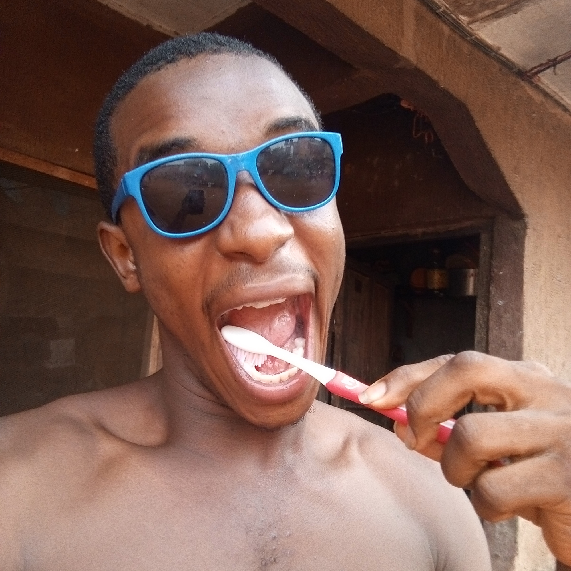 An undressed man with black hair and blue sunglasses is holding a pink toothbrush inside his open mouth, as though he is brushing his teeth. The man is in front of a building in the city of Onitsha, Nigeria. The building has brown wood walls and a shelf inside with kitchen pots and a yellow container.