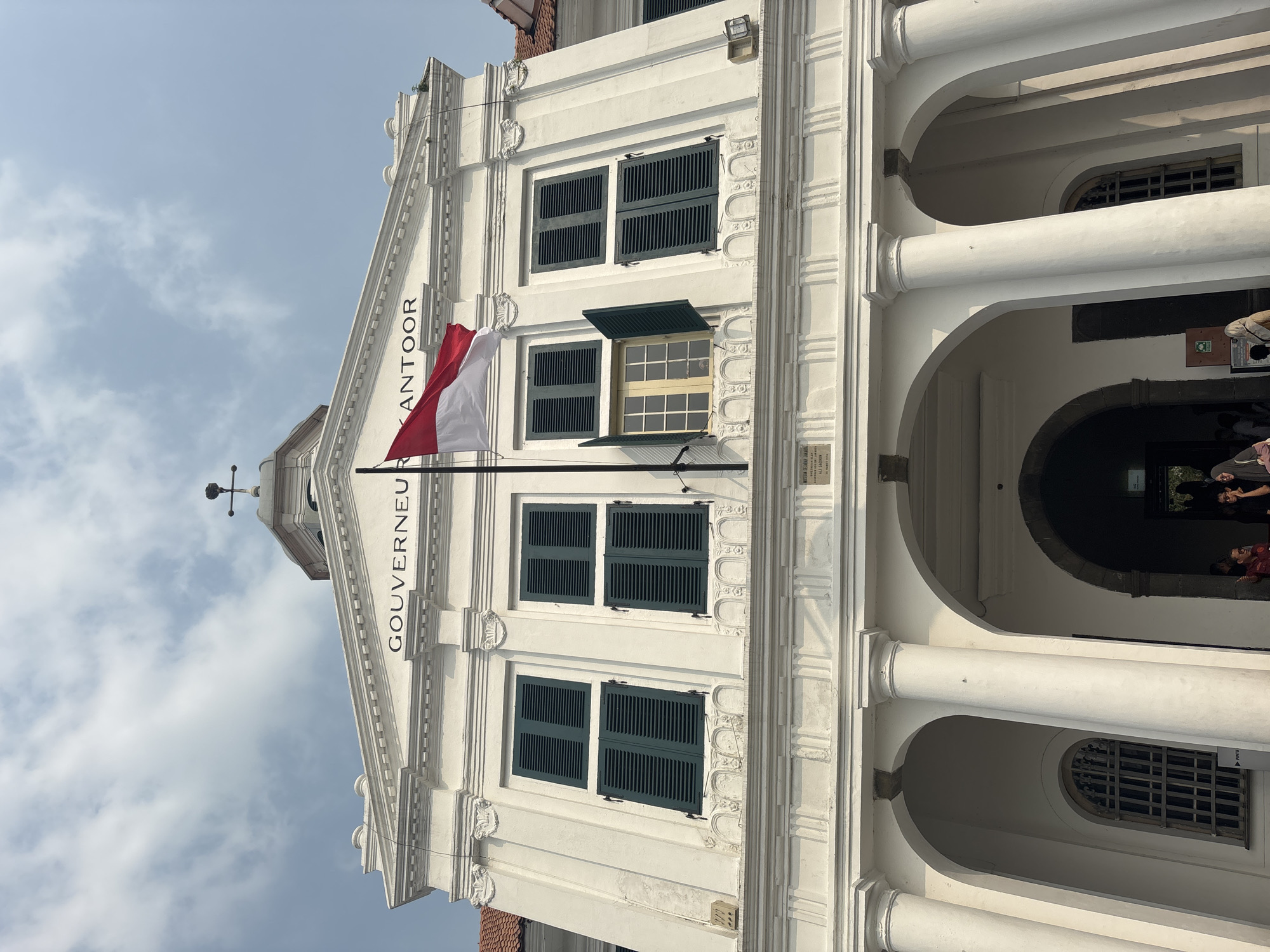 The image shows the exterior of the Fatahillah Square, also known as the Jakarta Old Town, in West Jakarta, Indonesia. The building features a grand white facade with arched doorways and columns. Above the entrance, the words "GOUVERNEUR KANTOOR" are inscribed in black lettering. The Indonesian flag, a horizontal bicolor of red and white, flies from a flagpole in front of the building. Several windows with dark green shutters are visible on the upper level, with one window partially open. The sky is partly cloudy. In the foreground, partially visible at the bottom of the image, are a few people.