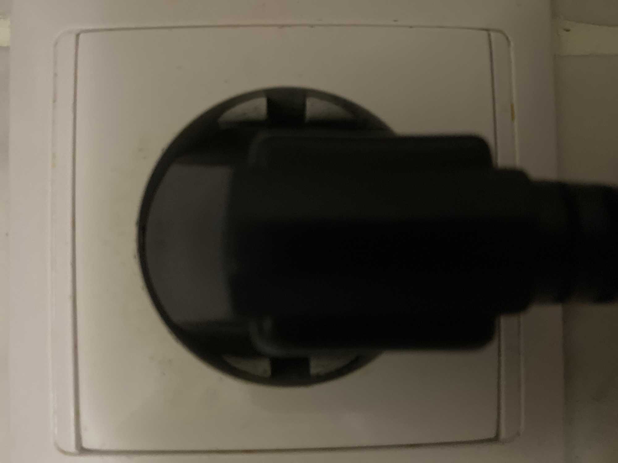 A black plug is inserted into a white electrical socket. The socket is part of a white wall outlet with a rectangular cover. The outlet has three circular openings. The plug is a dark color and obscures the center of the socket. The image is taken at a close-up, head-on angle. The background is a neutral, off-white wall. There is no visible indication of the location of the city.