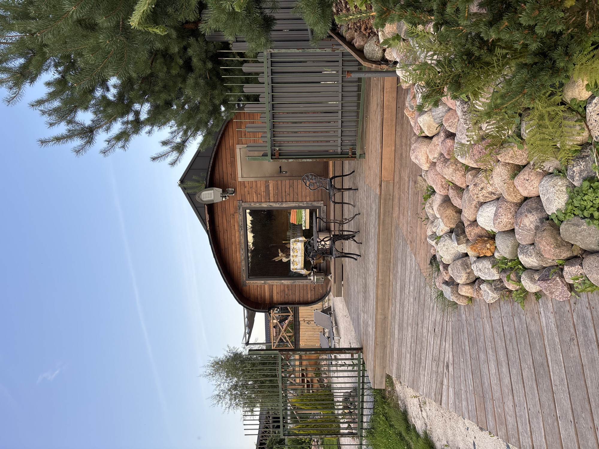 The image depicts a picturesque outdoor scene at a modern barrel sauna facility located in Zvenigorod, Russia. The central focus is a barrel-shaped wooden sauna building with a large panoramic window showcasing an interior setting with a table, chairs, and decorative items. In front of the sauna, a wooden deck area features a small wrought-iron table and two chairs. The deck is bordered by a rocky landscape with a variety of large, rounded stones and lush green foliage.
To the left of the deck, a wooden pathway made of planks leads away from the sauna and toward a metal fence. Beyond the fence, more vegetation and what appears to be a smaller structure or garden feature are visible. A tall pine tree dominates the right side of the frame, its branches extending over the scene. The sky above is a clear, pale blue with faint wisps of clouds, suggesting a calm, possibly late afternoon or early evening atmosphere. The overall impression is one of a serene and well-maintained recreational space.