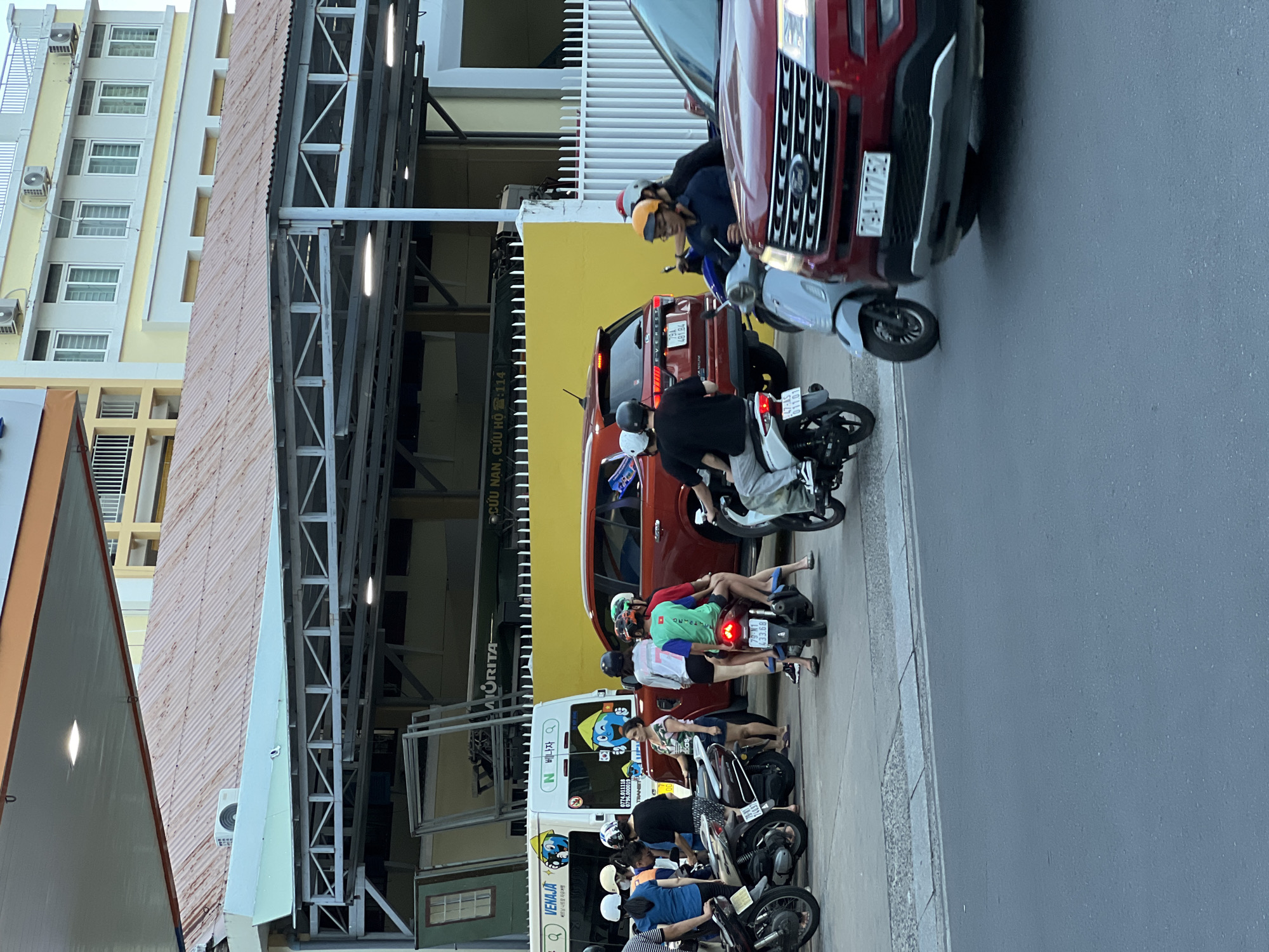 The image depicts a street scene in Nha Trang, Vietnam, likely near a gas station. Several individuals are on or near motorcycles. In the foreground, a red Ford Explorer is parked, with its front bumper visible. To the left of the Explorer, a light blue scooter is parked. Behind the scooter, a person in a black shirt and helmet is riding a white motorcycle. A red SUV is parked behind the white motorcycle. Further left, a group of people are gathered near a white van with Korean text and a cartoon logo. Several motorcycles are parked around the van, and people are standing next to them. Two individuals are seated on a black motorcycle in the middle ground, with one wearing a green shirt and the other a red shirt. A red SUV is parked behind them. The background shows a building with a corrugated metal roof. The overall scene suggests a busy urban environment with significant motorcycle traffic.