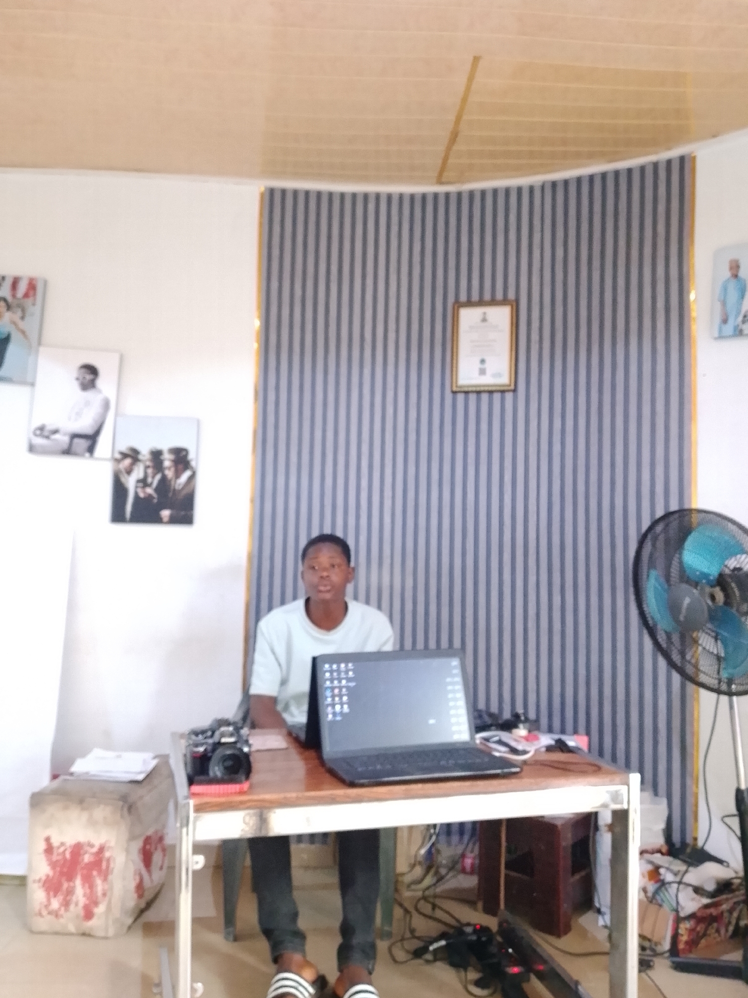 A young man in a light blue t-shirt and dark pants is seated at a wooden desk in a room with striped wallpaper. He is looking forward and appears to be speaking. In front of him on the desk is an open laptop, a camera, and some papers. To the left of the desk is a rectangular object with red markings. To the right of the desk, a black electric fan is visible. The room has a light-colored ceiling with horizontal lines. On the wall behind the man, there are several framed pictures. A ceiling fan is also visible. The setting is likely an indoor space in the city of Jos, Nigeria, possibly an office or a studio.