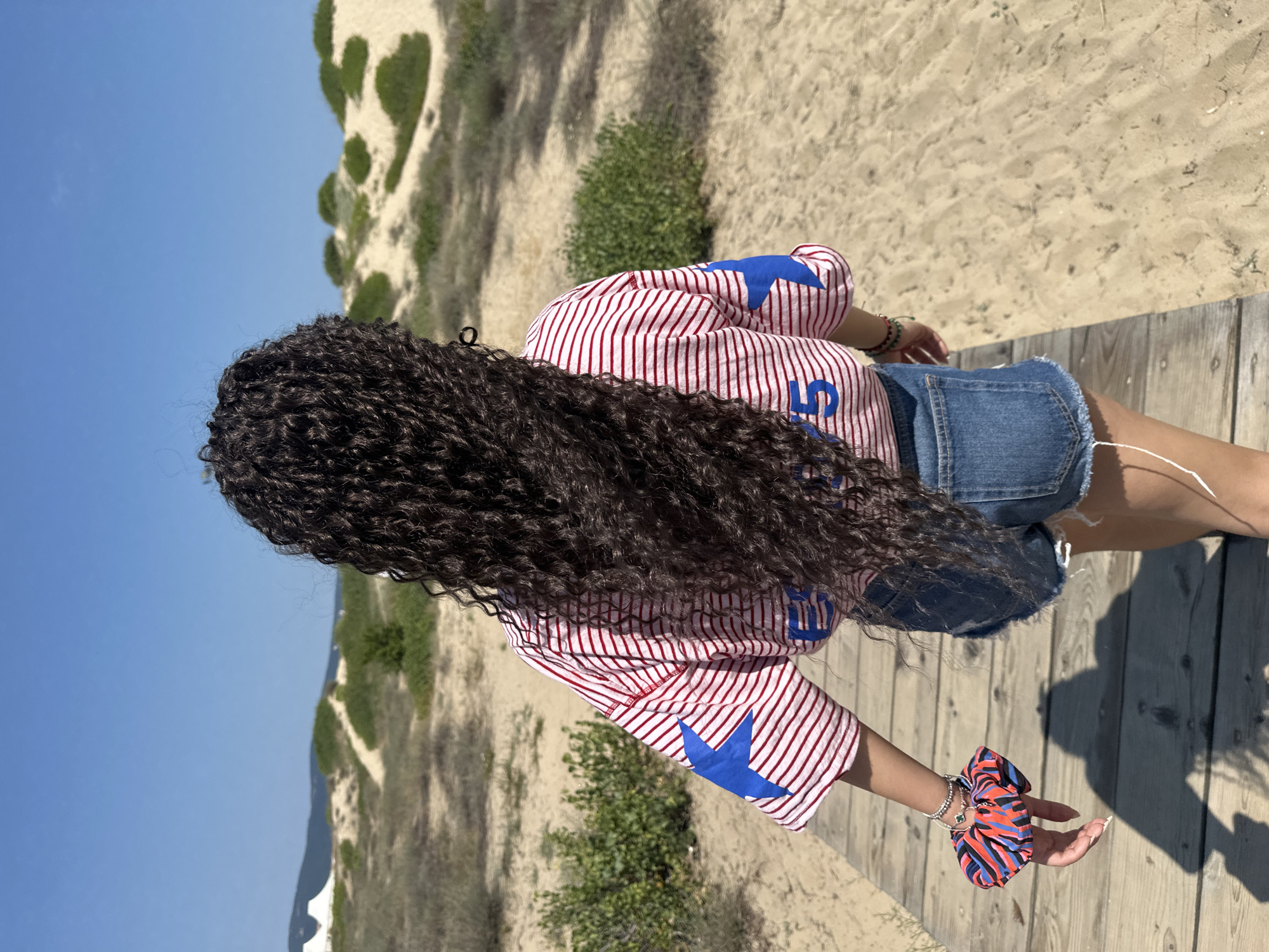 A young woman walks on a wooden boardwalk on a sunny day. She is viewed from behind, with her long, dark, curly hair flowing down her back. She wears a red and white striped t-shirt with large blue stars on the sleeves and a blue number "5" on the back. She also wears denim shorts. On her left wrist, she has a multi-colored scrunchie and bracelets. The boardwalk is situated on a sandy dune area with sparse green vegetation. In the background, there are more sand dunes and a clear blue sky. The location is the area of sand dunes in Nesebar, Bulgaria, a place known for its archaeological and historical significance.