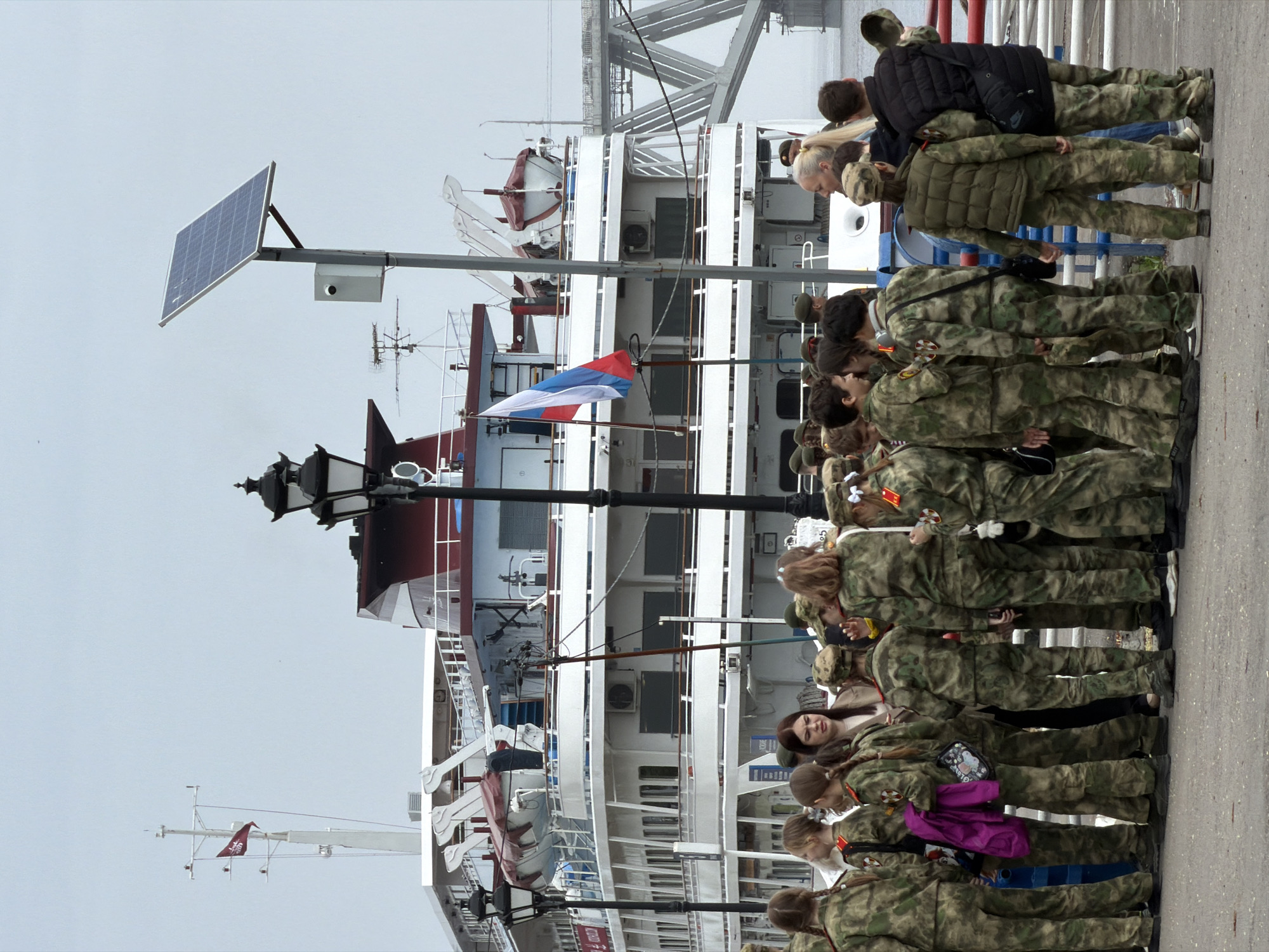 A sideways wide shot shows a group of people standing near a white boat. The people are wearing similar camouflage outfits. The boat has a red, white, and blue flag on its mast. The sky is overcast and gray. A black street lamp is to the left of the boat. A solar panel is mounted on a pole to the left of the street lamp.
Based on the architectural style of the boat and the presence of the Russian flag, it is likely that the location is in Russia. The city of Saratov is a major port city on the Volga River, and this is likely the location depicted in the image.