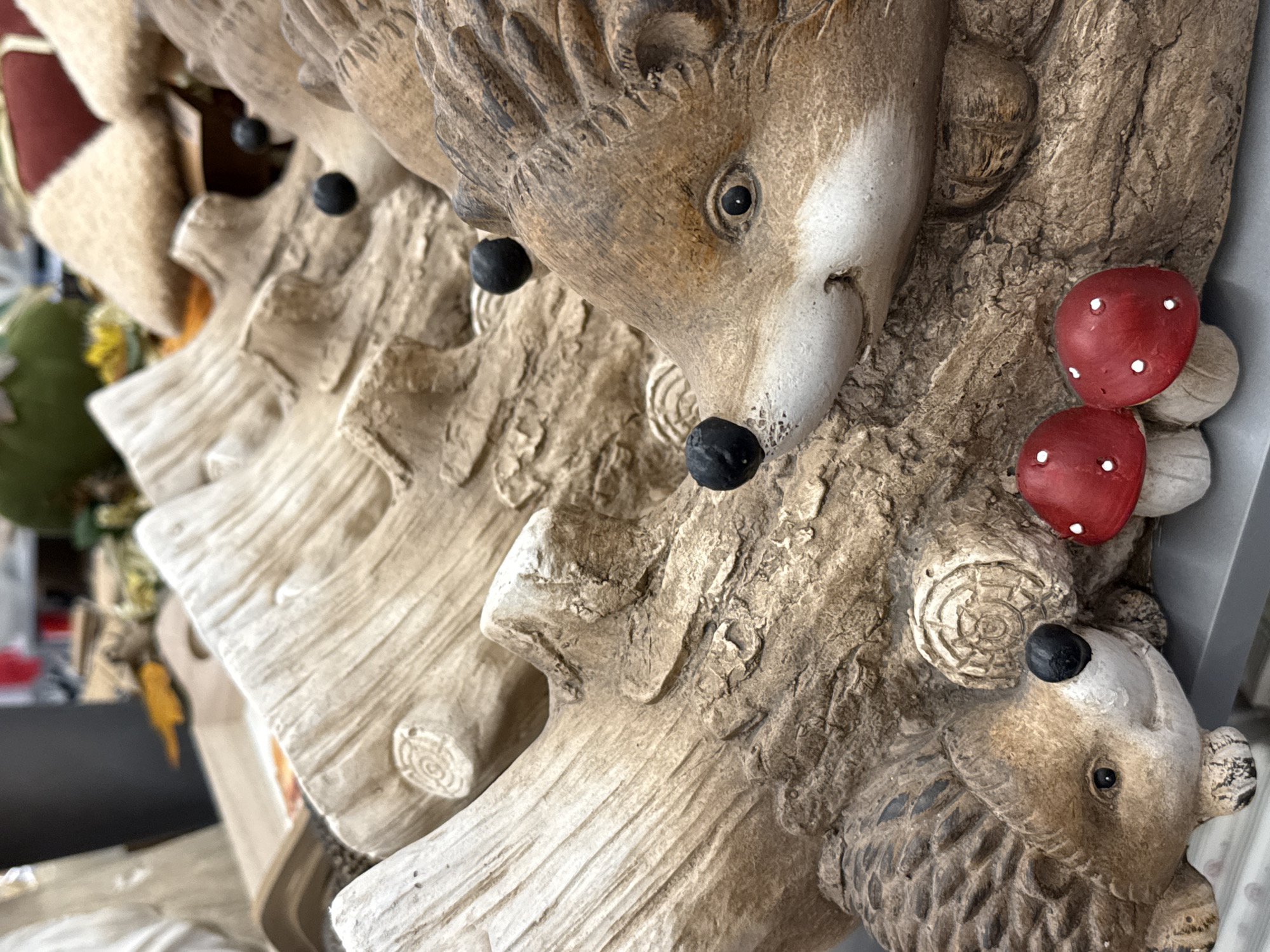 The image is a close-up, eye-level shot of a decorative garden display featuring two ceramic hedgehogs nestled among artificial logs. The hedgehogs, rendered in earthy brown tones with white snouts and black eyes and noses, are positioned as if emerging from the textured logs. One hedgehog is in the foreground, facing right, with its head angled slightly upwards. The second hedgehog is partially visible in the lower left of the frame, facing towards the right as well. The logs themselves have a rough, weathered texture and are carved to resemble natural wood, complete with circular knot details. Two small, red ceramic mushrooms with white spots are placed in front of the hedgehogs, adding a touch of whimsy to the scene. The background is blurred, showing hints of other decorative items and a neutral-colored surface, suggesting this display is located in a retail environment or a garden center. The lighting is even and diffused, highlighting the details of the ceramic figures. The overall impression is one of a charming, autumnal garden decoration. The scene is likely located in the city of Bad Lauterberg im Harz, Germany, given the context of the provided location.
