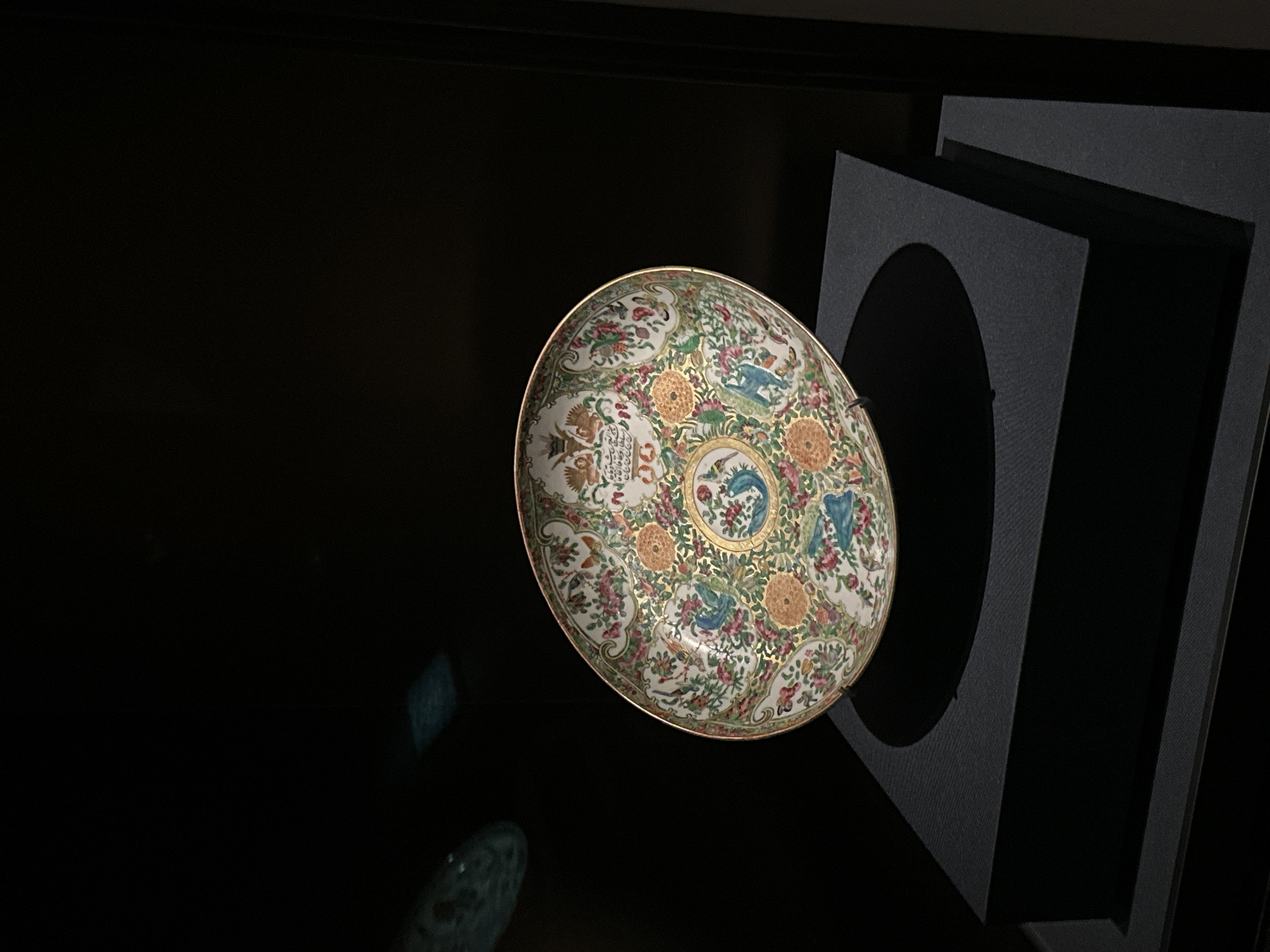 A decorative porcelain plate with intricate floral and bird motifs, accented with gold trim, is displayed on a black pedestal within a dimly lit exhibition space. The plate rests on a dark, textured surface, casting a distinct shadow. The background is predominantly black, with subtle reflections of what appear to be other display elements or lights visible in the upper left portion of the image. The overall presentation suggests a museum or gallery setting.