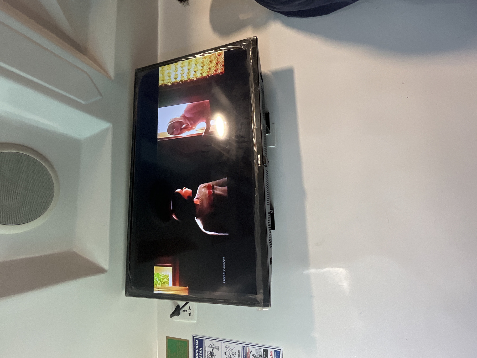 The image shows a television screen displaying a scene with two individuals. On the left side of the screen, a person with short dark hair is facing away from the viewer, with their back partially visible. They appear to be wearing a light-colored top with some red design. To the right, another person is visible from the chest up, wearing a pink hooded jacket and looking towards a window. The television is mounted on a white wall. Above the television, the ceiling has an interesting architectural design with recessed lighting. To the left of the television, a white electrical outlet with a black plug is visible, and next to it, a green sign with text in Indonesian. The overall setting appears to be indoors, likely a room or a public space. The presence of the Indonesian text on the sign suggests the location could be Indonesia, possibly in a public transportation hub or establishment in Karimunjawa, an island known for its beaches and marine tourism.