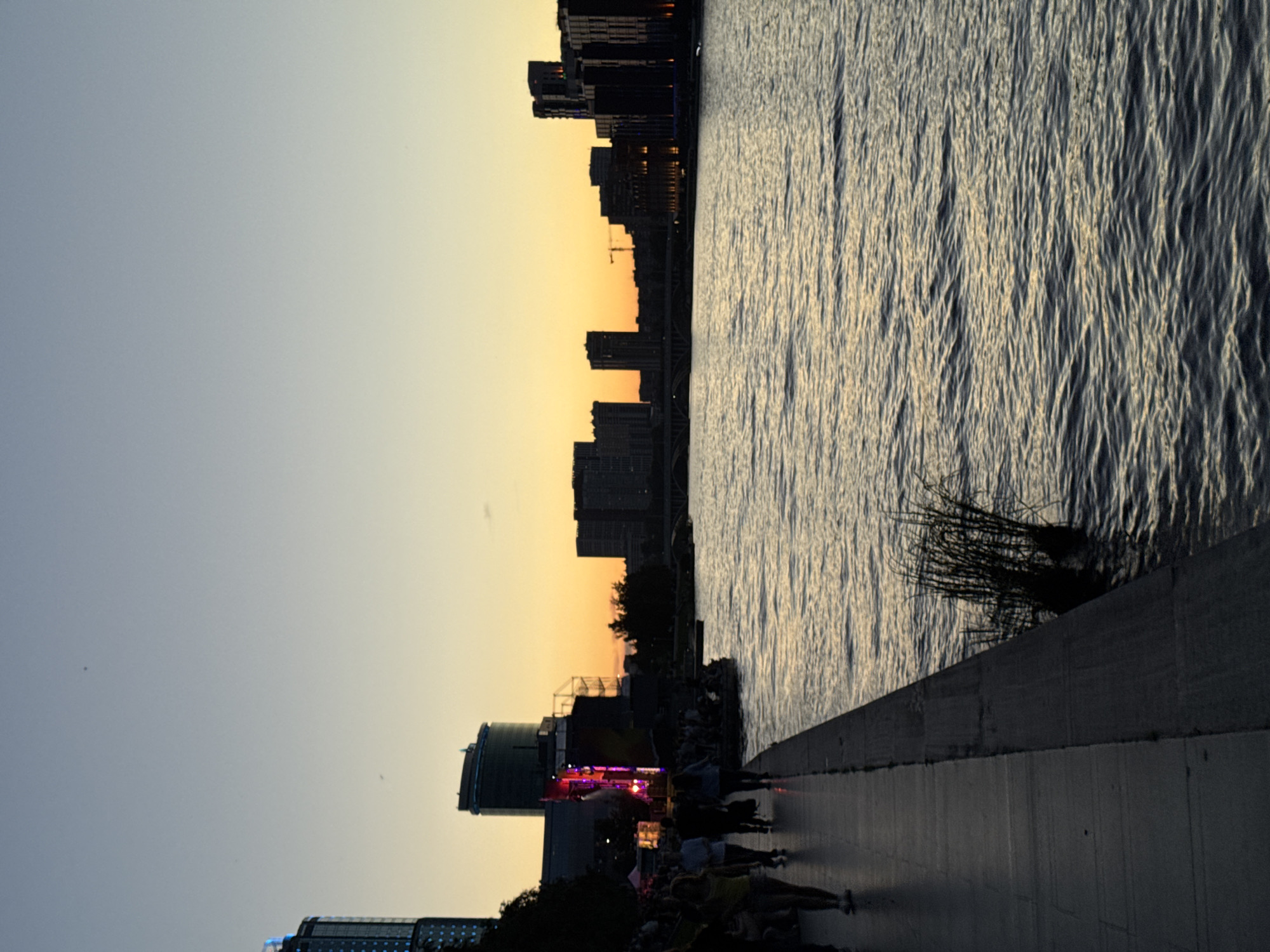 The image depicts a waterfront promenade in Yekaterinburg, Russia, at dusk. The sky transitions from a pale blue overhead to a soft orange and yellow gradient near the horizon, indicating the setting sun. Silhouetted against the sky are several modern buildings forming a city skyline, with some structures illuminated. In the foreground, a paved walkway runs along the edge of a wide body of water, likely the Iset River. Several people are walking along the promenade, their forms silhouetted in the dim light. A cluster of tall reeds grows at the water's edge on the right side of the image. The water itself reflects the colors of the sky and appears rippled. The overall atmosphere is calm and serene.