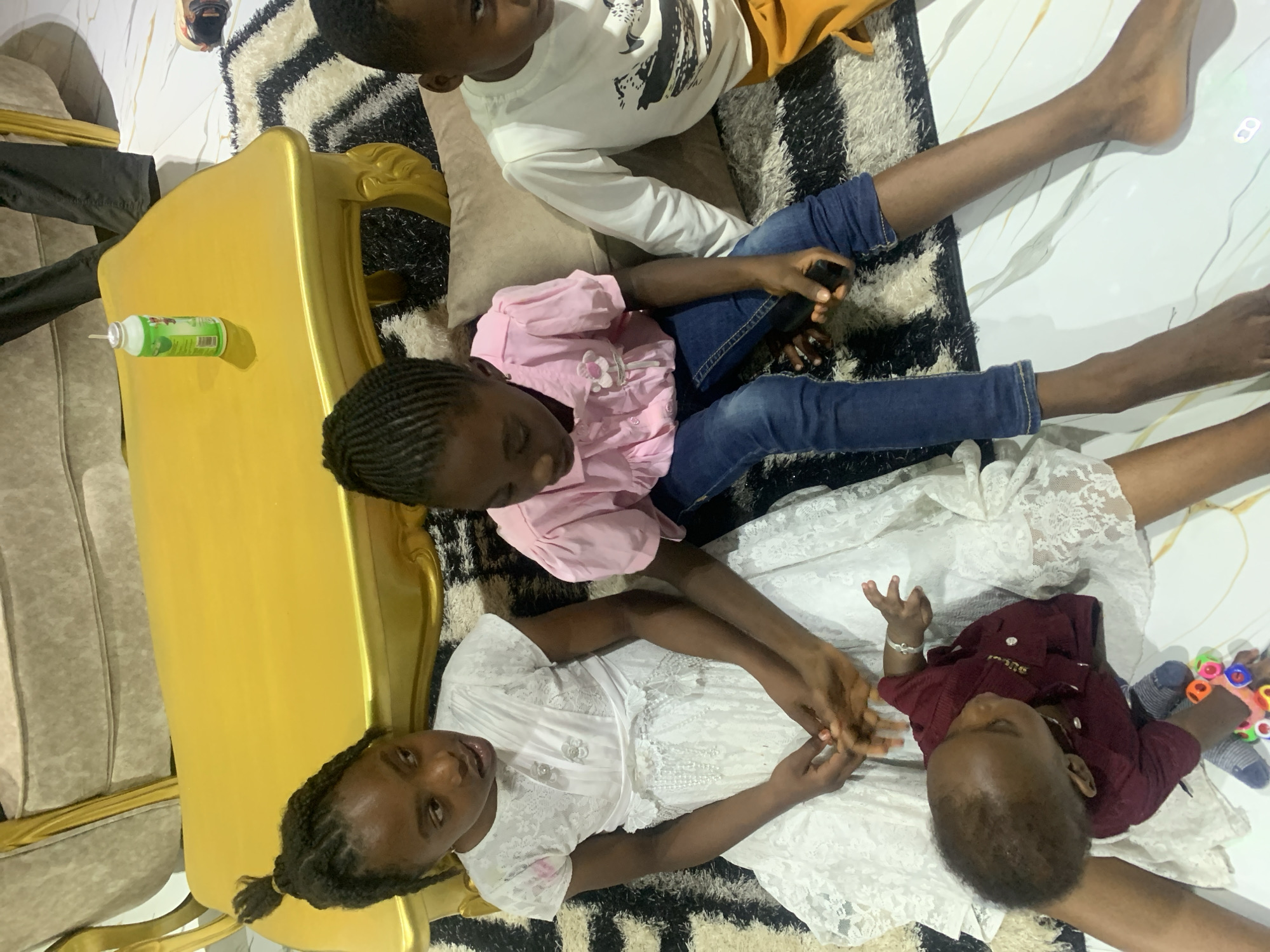 The image depicts four children indoors, possibly in a home in Jos, Nigeria. A child wearing a white long-sleeved shirt and mustard-colored shorts sits next to another child in a pink long-sleeved shirt and blue jeans. Another child wearing a white lace dress is near an infant in a maroon shirt. The children are on a rug with a black and white striped pattern. A gold-colored sofa is visible to the left, with a can of insecticide on its arm. The children are engaged in interactions, with the child in the white dress holding the hands of the infant. The environment includes a white, shiny floor beyond the rug.