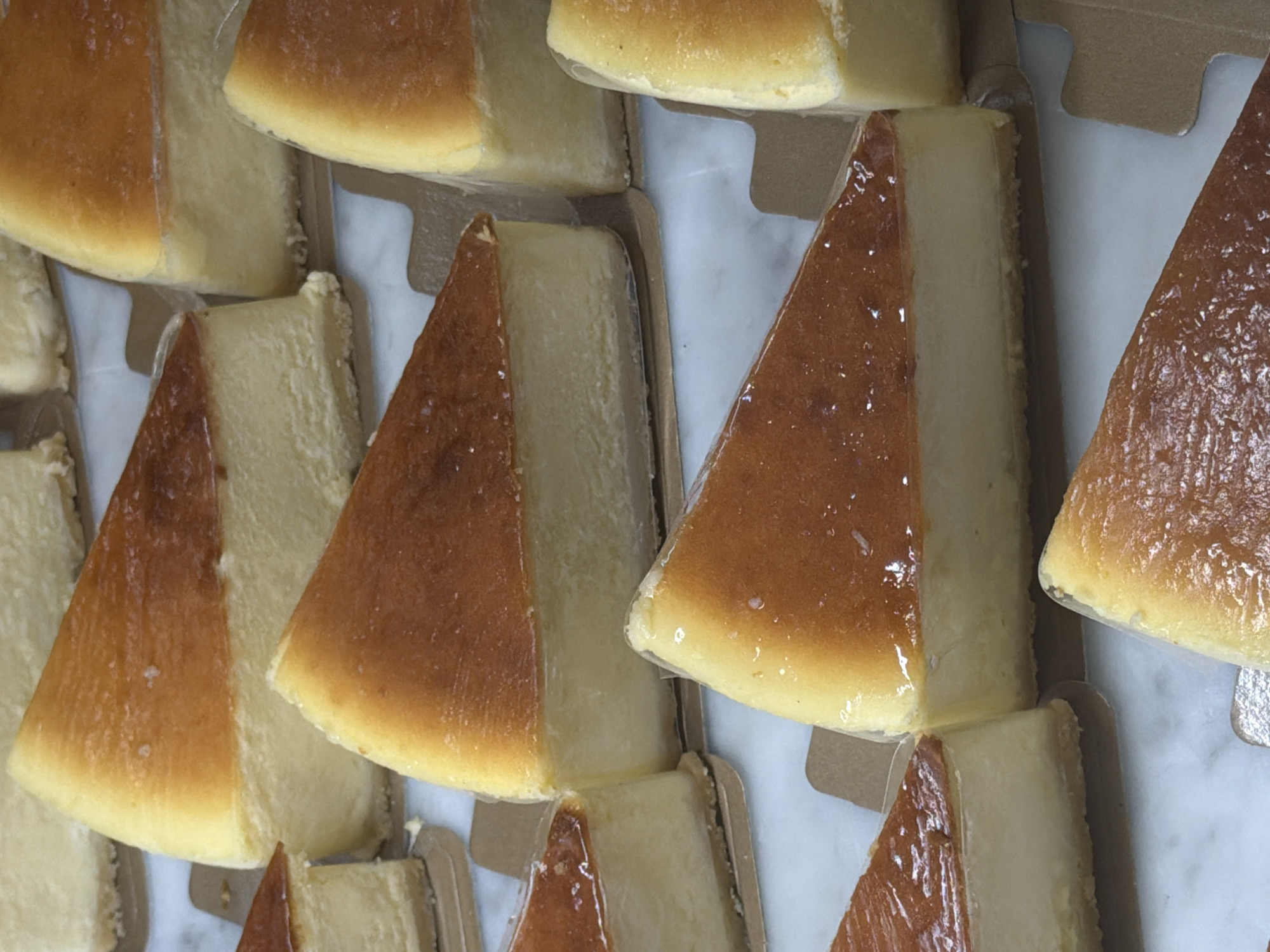 The image presents a close-up, high-angle view of numerous individual slices of cheesecake. Each slice is triangular, displaying a distinct two-layered appearance: a glossy, golden-brown top layer that appears caramelized or baked, and a lighter, creamy yellow bottom layer. The texture of the top layer shows subtle irregularities. All slices are individually encased in clear plastic wrapping, which creates slight reflections. They are presented upright in light brown, individual cardboard holders or trays. These holders are arranged in a dense pattern, suggesting a display or packaging for sale. The background is a light-colored, possibly white or marble-patterned, surface. No people are visible, nor is any specific activity taking place beyond the static arrangement of the desserts. The setup implies a commercial environment, such as a bakery, cafe, or market stall in North Kuta, Indonesia. There is no visible text within the image. The time of day and weather conditions cannot be determined.
