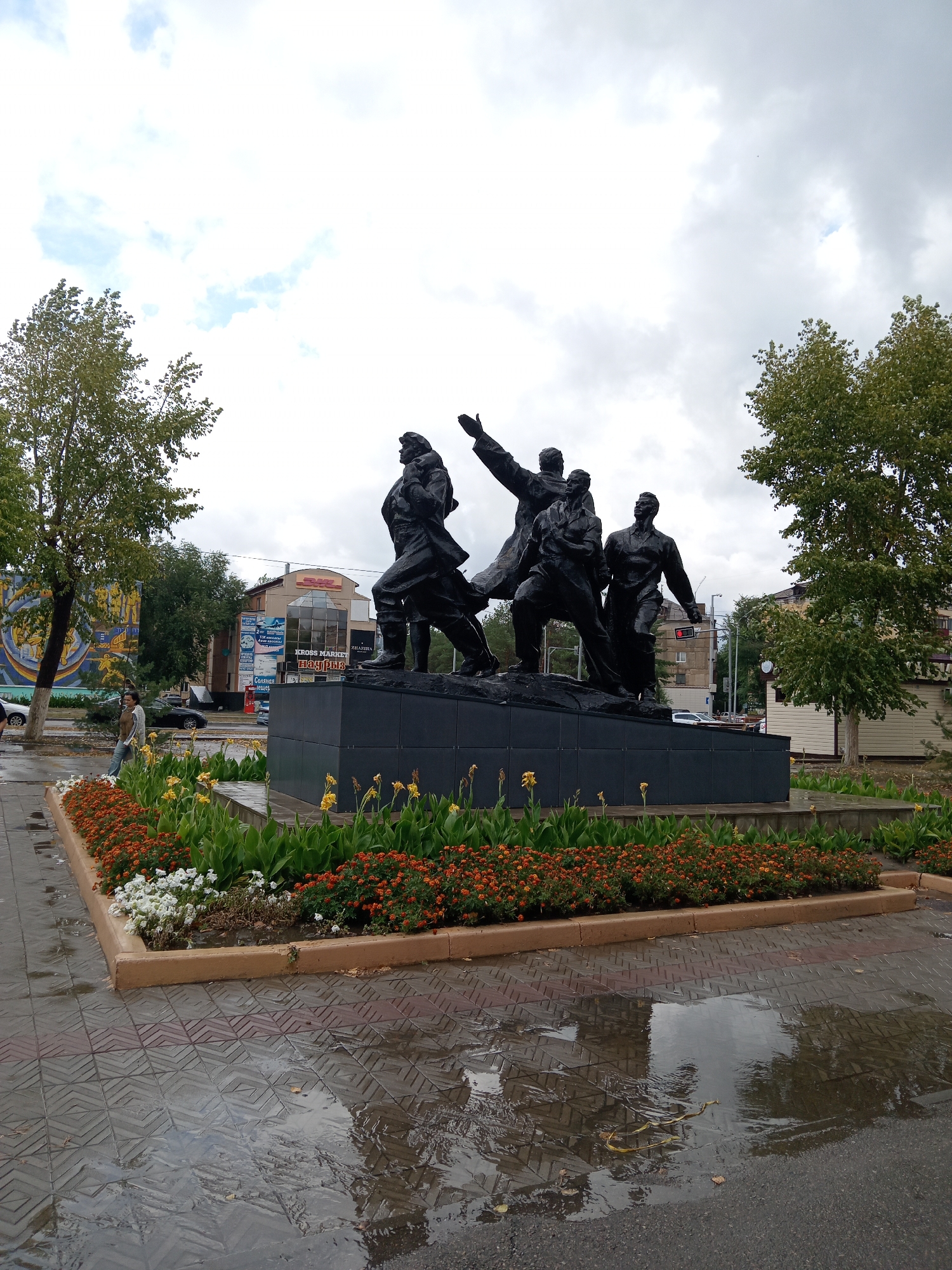 The image captures a bronze statue of four figures in dynamic poses, possibly representing a historical or ideological theme, situated in Rudny, Kazakhstan. The monument stands on a dark, rectangular pedestal, surrounded by a planter box filled with dense, colorful flowers, including orange, yellow, and white varieties. Beyond the monument, a street scene unfolds with several buildings visible, including one with a DHL logo. The ground in front of the monument is paved with patterned tiles, and due to recent rain, there are significant puddles reflecting the sky and surrounding elements. Two trees frame the monument, one on the left with vibrant green foliage and another on the right, also with green leaves. A lone woman, dressed in a light-colored top and dark pants, walks on the sidewalk to the left of the statue. Cars are parked and driving on the street behind the planter, indicating an urban setting. The sky is overcast with a mix of white and grey clouds. The overall impression is a public monument in an urban environment after a rain shower.