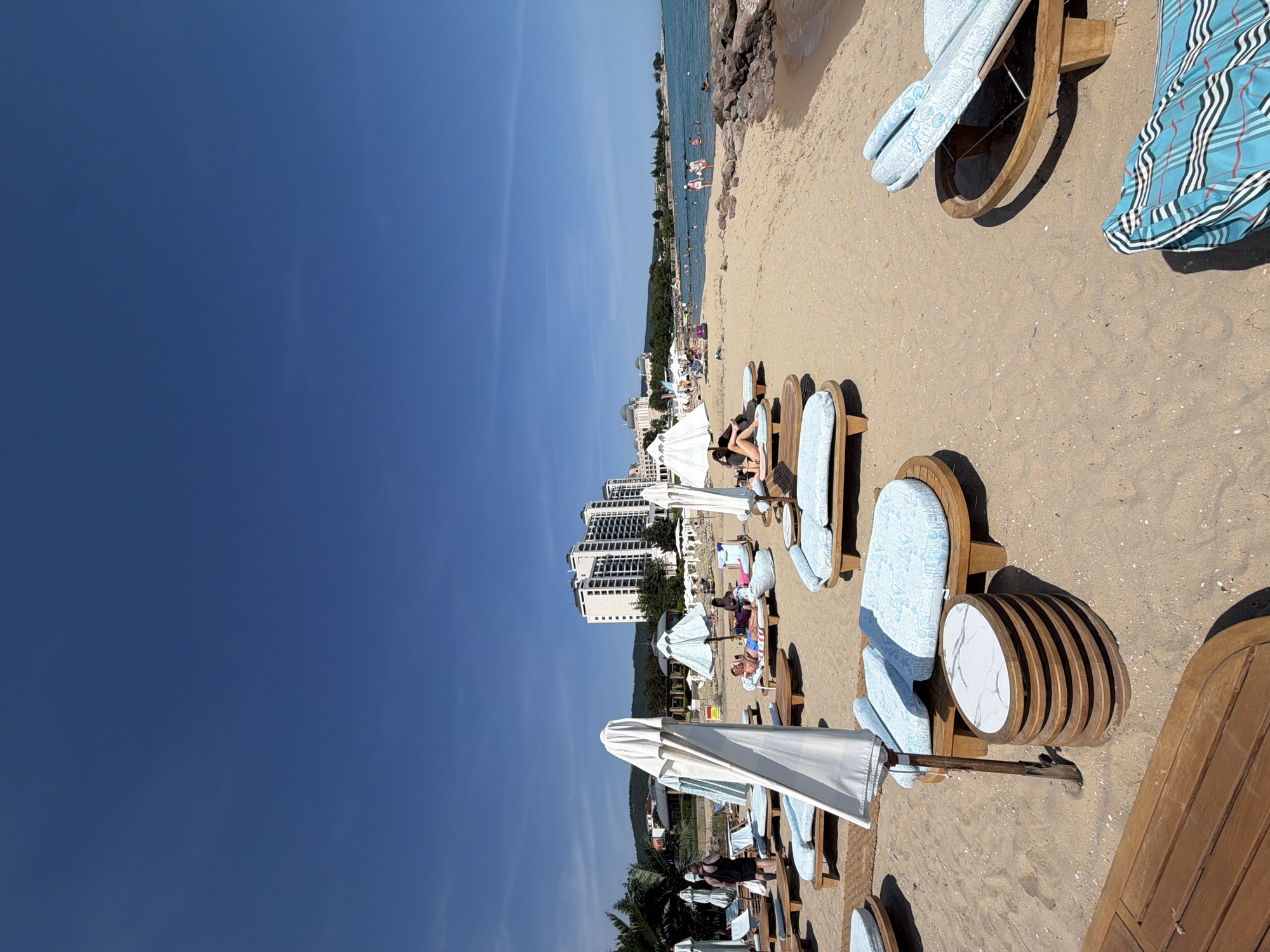 This image depicts a sunny beach scene in the city of Unknown, Unknown, under a clear, deep blue sky with minimal, faint wisps of white clouds visible in the upper right. The scene is dominated by a wide sandy beach extending into the distance, bordered by the calm, blue-green ocean on the right.
In the background, several multi-story modern white buildings, likely resorts or hotels, line the distant shoreline, with a large, lush green, tree-covered hill rising behind them on the left. The distant coastline curves to the right, where a cluster of dark, craggy rocks forms a natural break at the water's edge. Further down the beach and in the water, numerous people are visible, some swimming and others standing near the shore.
The midground and foreground of the beach are equipped with numerous wooden lounge chairs featuring light blue, patterned cushions. Many white beach umbrellas are scattered across the sand; some are open, providing shade, while others, like the prominent one in the left-center foreground, are closed and upright. A unique oval side table with a white marble-patterned top and a stacked wooden slatted base is positioned next to a closed umbrella in the mid-foreground. In the bottom right corner, a light blue and white striped blanket with a distinctive pattern is draped over a lounge chair.
Several individuals are present on the beach closer to the viewer. A man in blue swim trunks lies on his back on a lounge chair in the mid-left. Another person is partially visible lying face down on a chair further to the right. On the far left, a person in dark clothing stands near a closed umbrella. The bright sunlight casts distinct shadows from the umbrellas and chairs onto the sand, indicating a high sun angle.