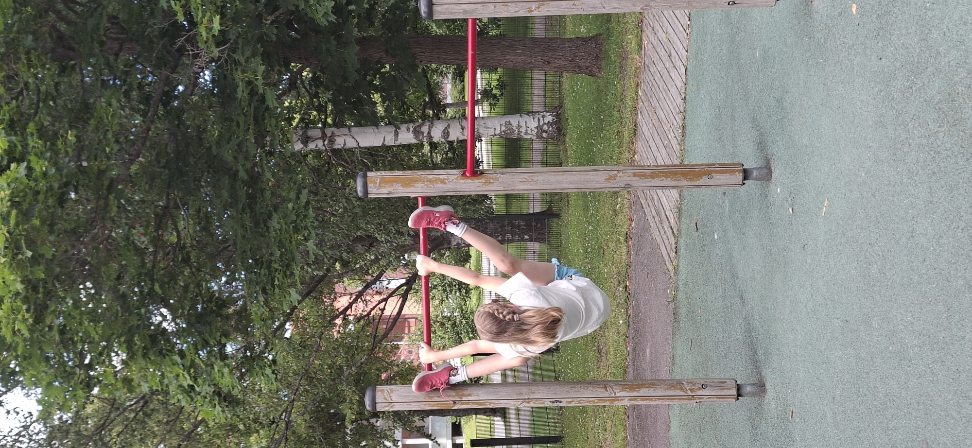 The image shows a young girl with braided blonde hair hanging from a horizontal bar on an outdoor playground in Lahti, Finland. She is wearing a white t-shirt, blue shorts, white socks, and pink sneakers. The playground has wooden beams and red metal poles. The ground is covered with a green mat, and a wooden walkway is visible to the side. In the background, there are trees with green leaves, a black metal fence, and a glimpse of a building with red brick. The lighting suggests it is daytime, likely a sunny or partly cloudy day. The girl's posture and the setting indicate she is engaged in playful activity on the playground equipment. There is no visible text in the image.
