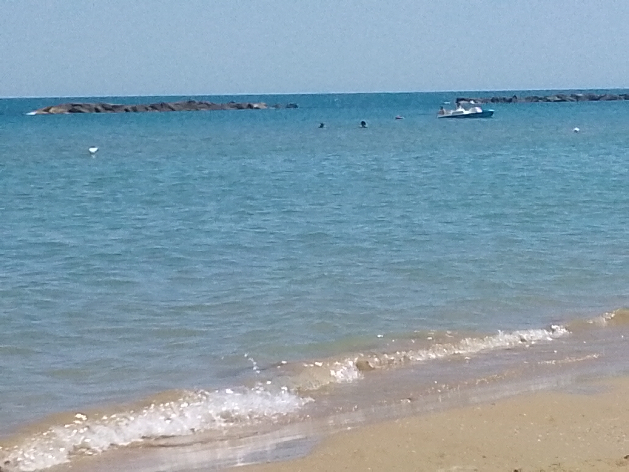 This is a wide shot of a beach on a sunny day. The ocean is calm with small waves lapping at the sandy shore. In the distance, there are two breakwaters made of large rocks. A small white boat with a blue trim is visible in the middle of the water, and a few people are swimming or floating nearby. The sky is a clear, light blue, suggesting fair weather. The image appears to be taken from the beach, looking out towards the sea. The time of day is likely midday or early afternoon due to the bright sunlight. There is no visible text in the image.