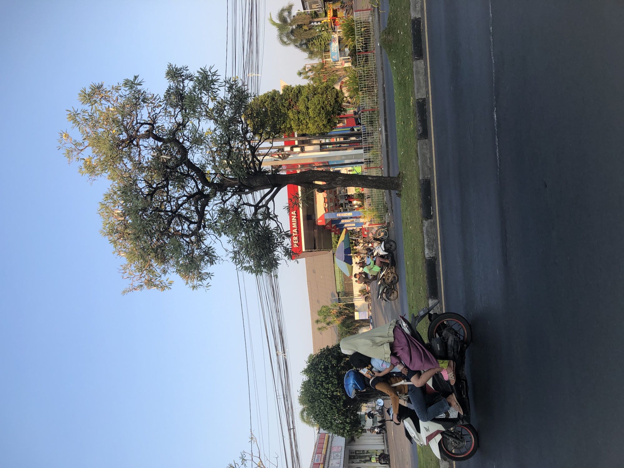 The image depicts a street scene in Gelam, Indonesia, with a Pertamina gas station prominent in the background. In the foreground, three individuals are riding on a white scooter. The person in the driver's seat wears a blue helmet and a brown jacket, with dark pants. The middle passenger wears a light blue shirt and appears to be holding a phone. The passenger in the rear wears a light green hijab and a long, dark purple dress. The scooter is traveling on a wet asphalt road. Behind the scooter, a grassy median separates the road from a sidewalk and the gas station. The gas station has red and white branding. Several other motorcycles and people are visible at the gas station and on the sidewalk. Trees are scattered throughout the scene, including a large tree in the center. Overhead, power lines crisscross the sky, which is clear and blue. The lighting suggests it is late afternoon or early evening.