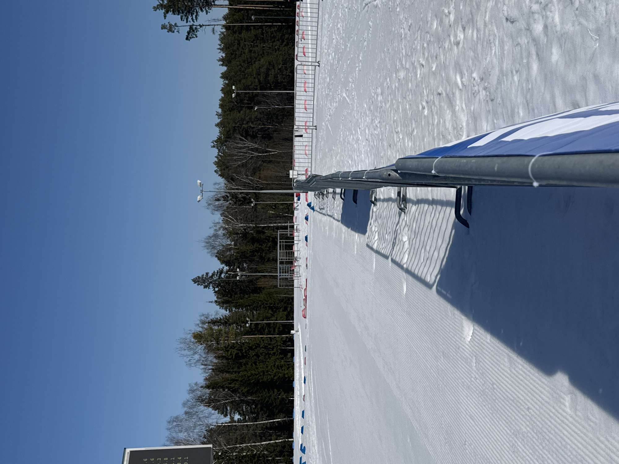 The image depicts an outdoor winter sports facility in Прикамский, Russia, under clear blue skies. The foreground and midground are covered in snow. To the left, a section of snow is groomed with parallel ridges, suggesting a track. To the right, the snow is less disturbed. A line of interconnected gray metal barriers extends from the bottom right towards the center-left, separating the two snow areas. A blue and white banner is affixed to the right side of these barriers. Long shadows of the barriers are cast onto the snow.
In the left midground, a large, dark electronic display board, partially visible, shows white numerical text. Scattered blue and red markers or cones are present on the snow surface in the midground. Further back, on the right side of the track, additional metal barriers are visible, along with some elevated, open-frame structures. The background features a dense line of coniferous and bare deciduous trees. Several tall light poles with multiple luminaires are strategically placed throughout the facility and along the treeline. No individuals or active events are present in the scene.