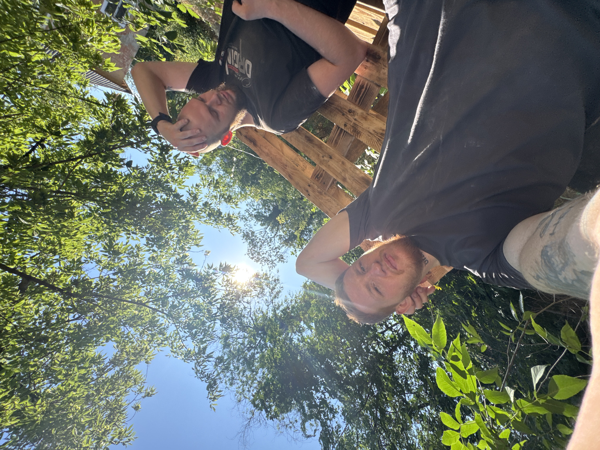 Two individuals are outdoors under a canopy of green leaves and a bright blue sky, with sunlight filtering through the foliage. The person in the foreground, a light-skinned male with ginger hair and a beard, is lying down with his left arm raised and hand behind his head, looking at the camera. He is wearing a dark t-shirt and has visible tattoos on his left arm. Behind him, to the right, another light-skinned male with a shaved head and a beard is also lying down, with his right arm raised and hand on his head, looking away from the camera. He is wearing a black t-shirt with white lettering. In the background, a partially constructed wooden structure, possibly made of pallets, is visible. The environment appears to be a natural setting with lush greenery and a hint of a building in the upper right corner. The perspective suggests the photo was taken from a low angle.