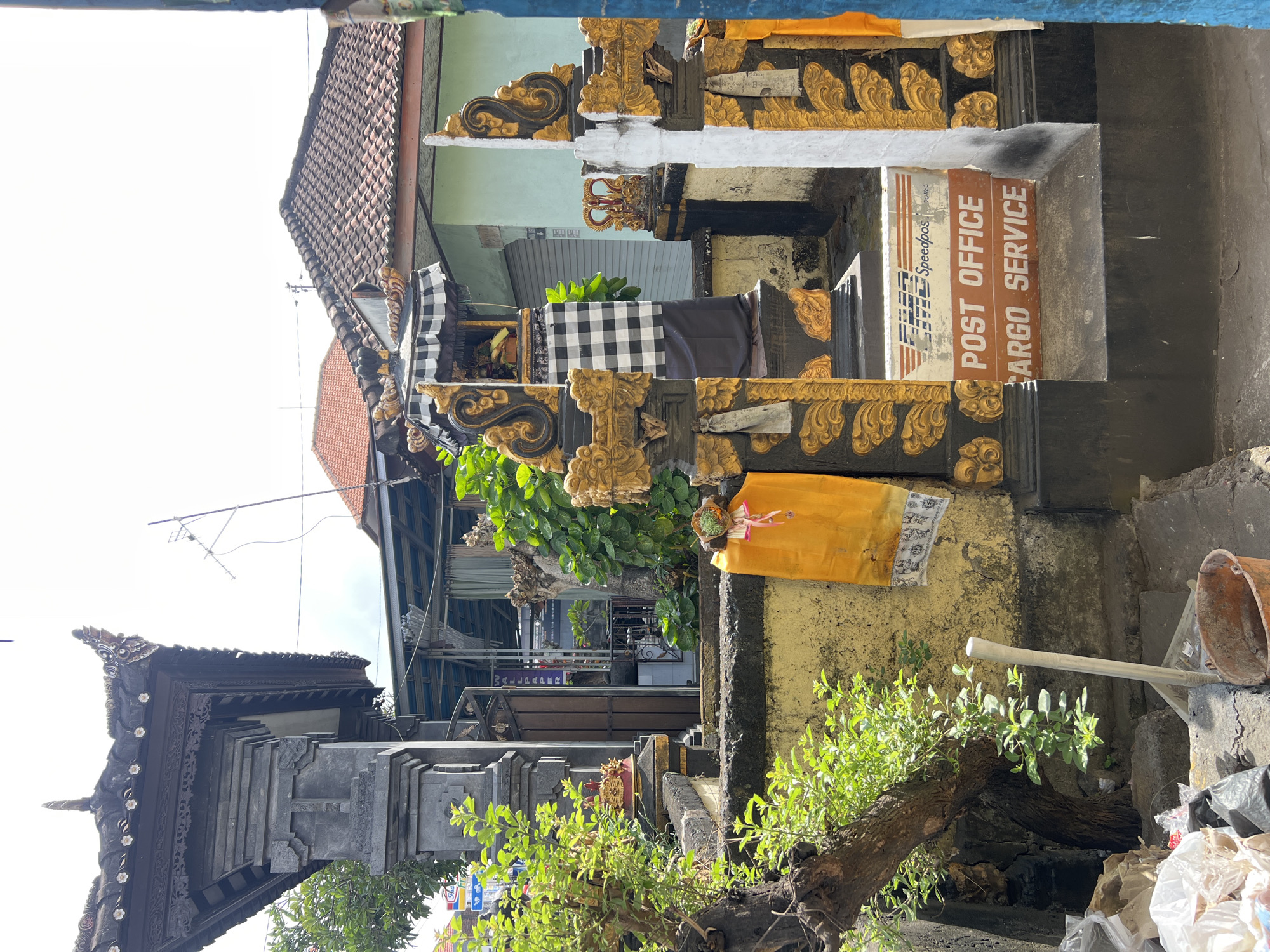 The image depicts an outdoor scene in Denpasar, Indonesia, with a focus on a traditional Balinese structure, possibly a temple entrance or shrine. The structure is made of dark stone and features intricate carvings. Adorning the structure are yellow and black checkered fabrics, typical of Balinese offerings. A prominent sign is visible, indicating "POST OFFICE" and "CARGO SERVICE" with a "Speedpost" logo, suggesting a postal service operating within or adjacent to this culturally significant site. To the left, a tree with lush green foliage grows, and to the right, a building with a tiled roof is partially visible. The ground in the foreground shows some debris, including plastic bags and a bucket. The lighting suggests it is daytime, with clear sky overhead. No people are explicitly visible in the image.