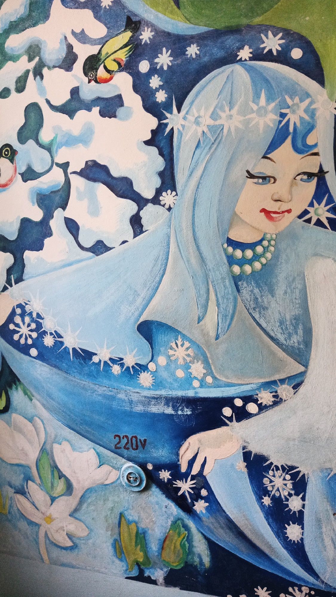 The image shows a close-up of a painting, likely a mural, depicting a winter scene with a young woman with long blue hair wearing a star-shaped crown and pearl necklace. She has blue eyes and red lips, and is dressed in white and blue garments. Snowflakes and stylized white flowers are also visible in the painting. In the background, there are abstract shapes in shades of blue and white, resembling snowdrifts or clouds, and two small birds, one yellow and black, the other red and black. The painting appears to be on an exterior surface, as a circular metal fixture with the inscription "220V" and a screw is visible in the lower center of the image. This fixture suggests the painting might be located on an electrical cabinet or a similar outdoor electrical installation. Based on the context provided, this artwork is located on the facade of a residential building at 10 Chornomorska Street in the city of Kryvyi Rih, Ukraine.
