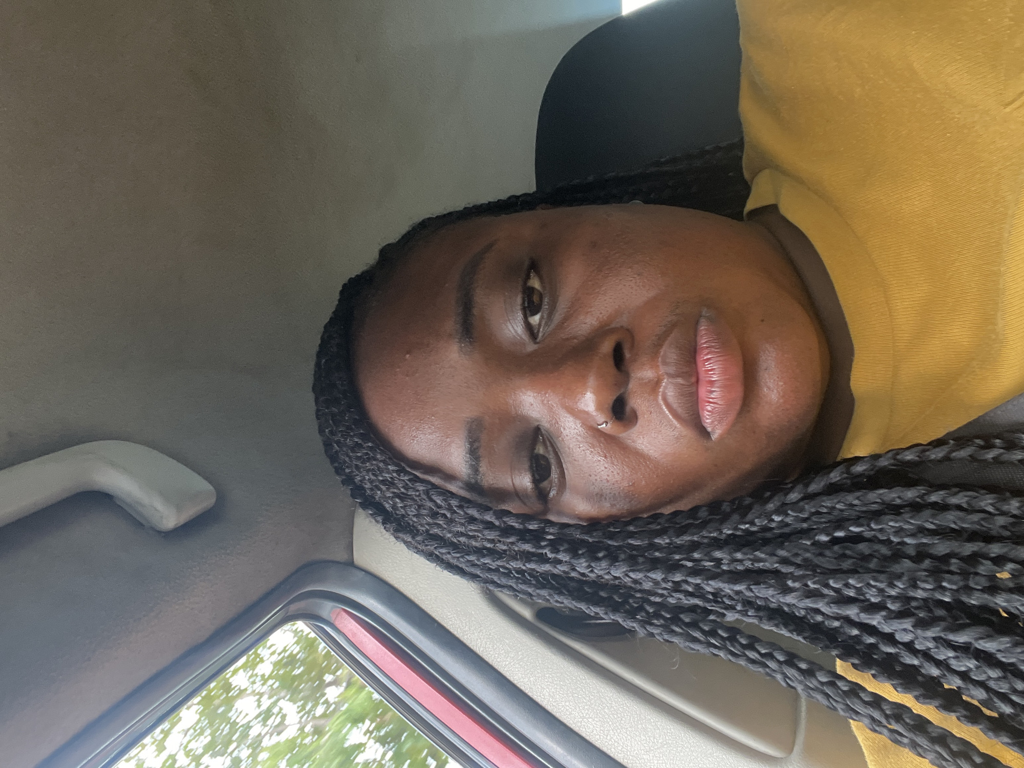 A close-up selfie captures a person of color, likely a woman, inside a car. The individual is positioned towards the left side of the frame, looking directly into the camera with a neutral expression. Their dark skin tone is prominent, and they have full lips and a small silver nose ring on their left nostril. Their hair is styled in long, dark box braids, cascading over their shoulder and chest.
The person is wearing a mustard yellow or golden yellow collared top, possibly a polo shirt, and a dark seatbelt is visible across their chest, suggesting they are a passenger or driver. The background reveals the car's interior, including a light-colored headliner and a grab handle on the left. A car window occupies the lower left portion of the image, showing blurred green foliage outside, indicating movement or a natural outdoor setting during the daytime. The lighting appears soft but bright, illuminating the person's face. No visible text is present in the image. Given the location context, this scene is set in Abuja, Nigeria.