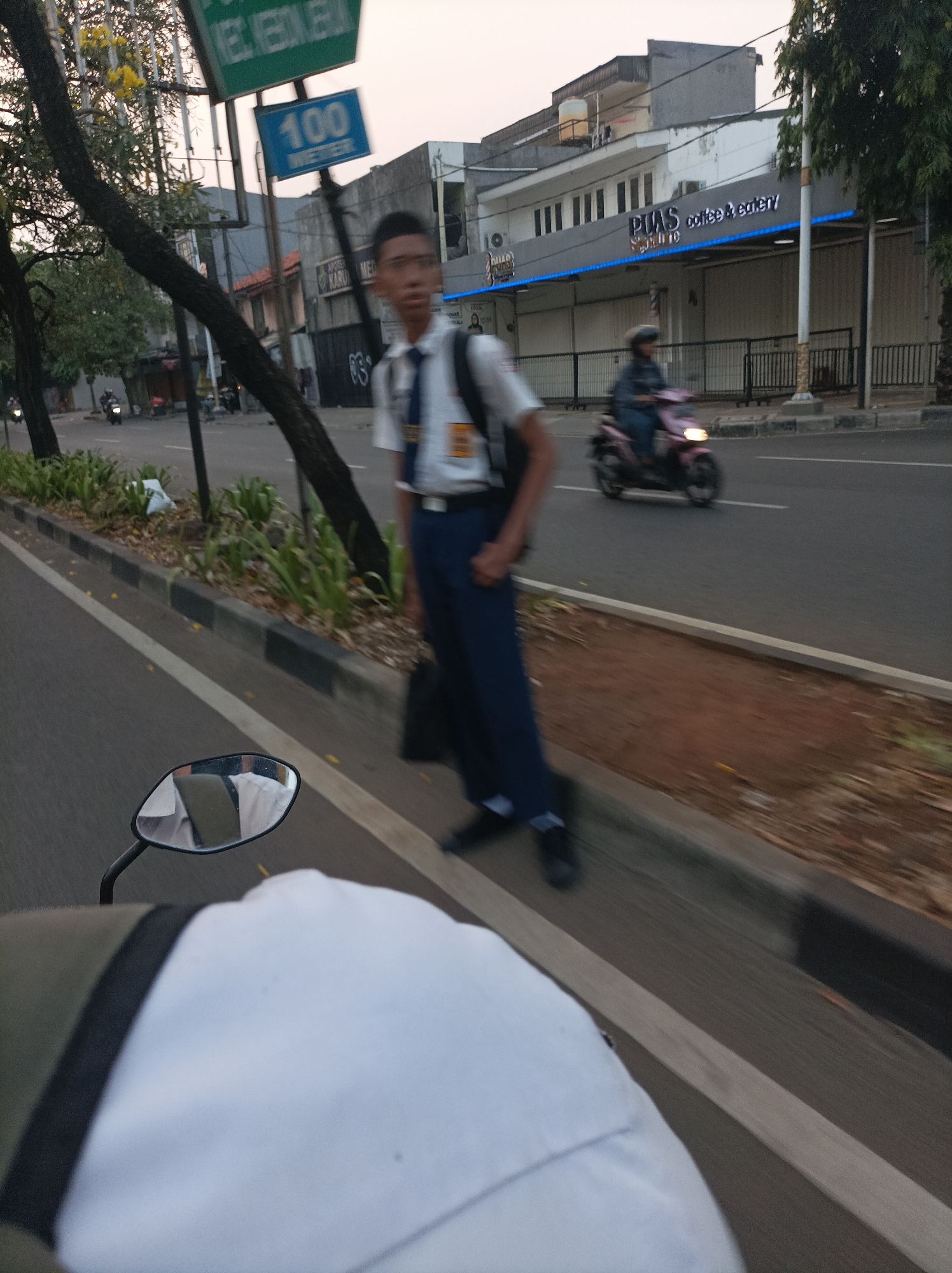 The image depicts a street scene in West Jakarta, Indonesia, likely during dusk. In the foreground, a person is riding on a motorcycle, evidenced by the visible rearview mirror reflecting the rider's white shirt and the blurred perspective of the road.
To the right of the road, a young male student wearing a white shirt, blue pants, and a school tie walks along the sidewalk. He has a backpack slung over his right shoulder. Behind him, a pink scooter carrying a rider passes on the road.
Further down the street, a building with the signage "PIAS coffee & eatery" is visible, illuminated by blue neon lights. The street is lined with trees and some greenery. Traffic signs are present, including one displaying "100 METER" in blue lettering. The sky suggests the sun is setting. The overall atmosphere is that of a typical urban environment in Indonesia.