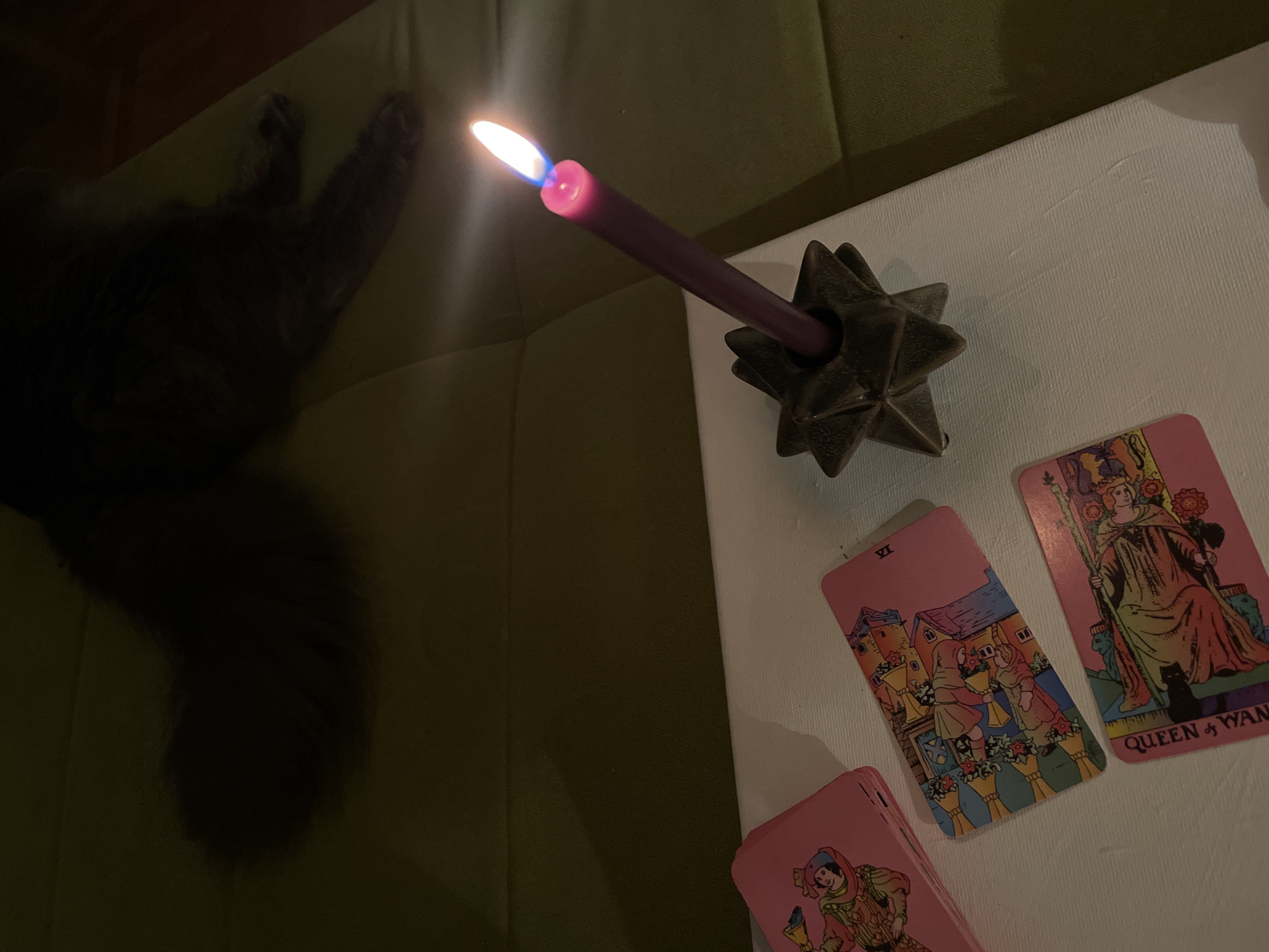 The scene depicts a dimly lit indoor setting, likely a domestic space. A dark-furred cat is partially visible on a green upholstered surface, possibly a couch or cushioned seat. Adjacent to this, on a white surface, likely a table or canvas, is a lit purple candle in a dark, star-shaped candle holder. Next to the candle are three tarot cards. One card is partially visible, revealing the bottom edge depicting a figure. The second card shows the "VI" card. The third tarot card is facing up, showing the "Queen of Wands" card. The city of Boreti, Montenegro is not discernible from the provided image. There are no people visible in the image.