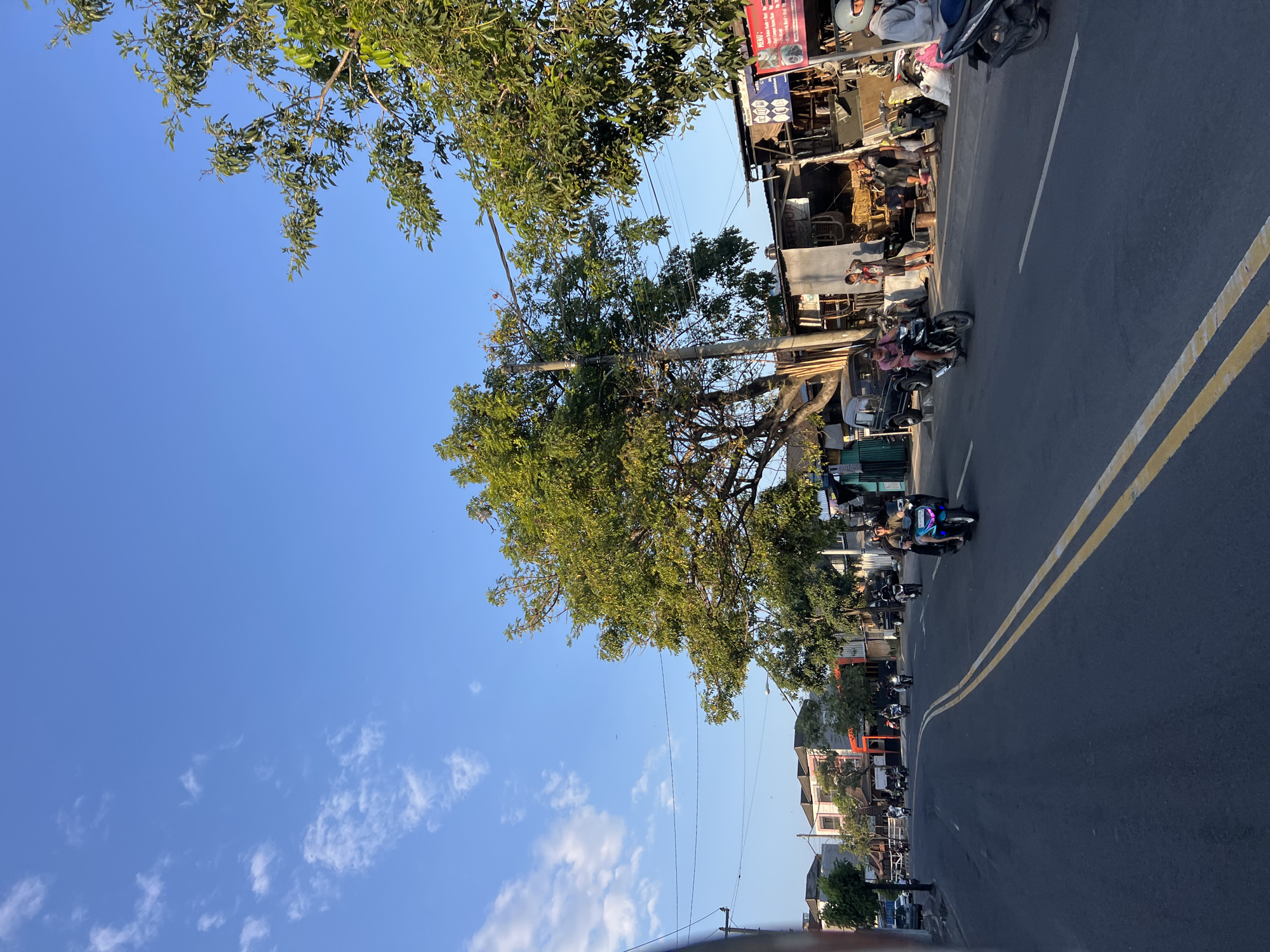 The image depicts a street scene in Denpasar, Indonesia, under a clear blue sky with scattered white clouds. The road is paved and has double yellow lines down the center. Several motorcycles are in motion on the road, with riders wearing helmets. On the right side of the road, there are shops and stalls with merchandise displayed, and some people are standing or sitting. A large tree with green foliage is prominent on the right side of the street, casting some shadow. Buildings line the left side of the road. The lighting suggests it is late afternoon or early morning.