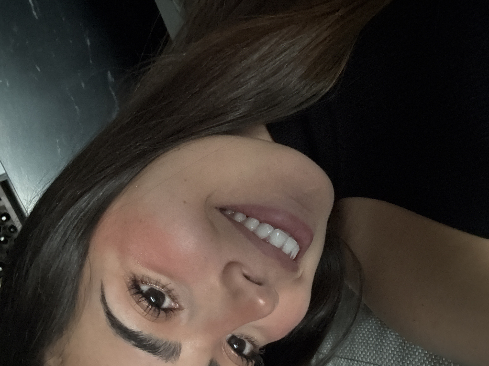 A close-up, angled portrait depicts an adult female positioned within an indoor environment in San Bernardino Tlaxcalancingo, Mexico. The subject has dark brown hair, fair to medium skin tone, prominent dark eyebrows, and dark eyes with visible eyelashes. Her cheeks exhibit a reddish flush. She is smiling, revealing uniformly white, unblemished teeth. She is dressed in a black high-necked top. The background on the upper left consists of a light-colored, textured fabric surface. The upper right background shows a dark, smooth surface with subtle white streaks, with indistinct dark, circular objects partially visible above it. The lower portion of the image includes a section of the textured fabric and the subject's black attire.
