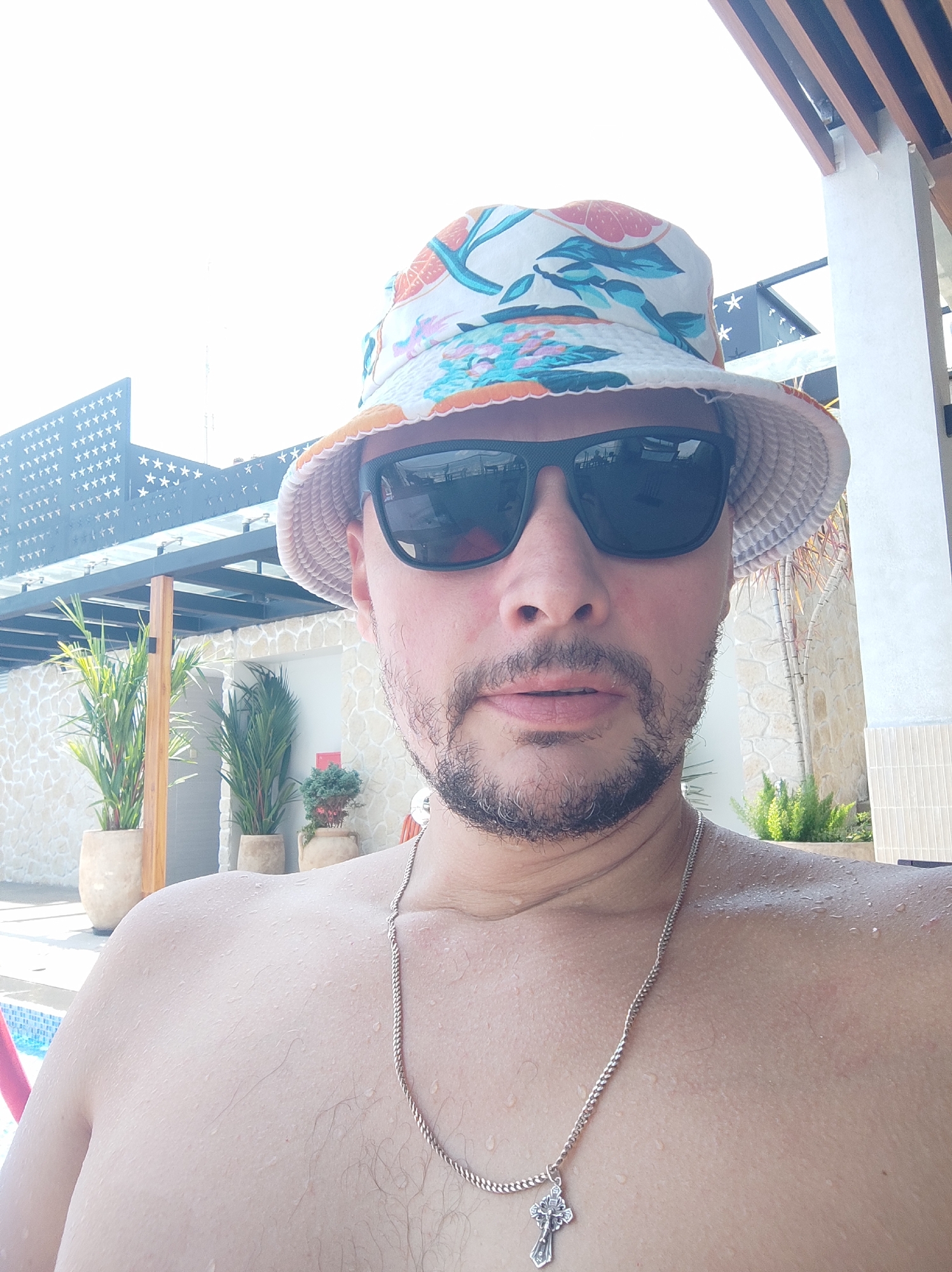 A man wearing a colorful bucket hat and black sunglasses takes a selfie outdoors. He has a beard and is shirtless, with visible water droplets on his skin, suggesting he has been in water. He is wearing a silver chain with a cross pendant. In the background, there is a modern building with a stone facade and wooden accents, as well as potted plants. The scene appears to be at a resort or a similar establishment, possibly near a swimming pool given the water droplets and the blue tiled edge visible in the lower left corner. The bright sunlight and clear sky suggest a warm, sunny day. The overall ambiance is casual and relaxed. This scene is set in Nha Trang, Vietnam.