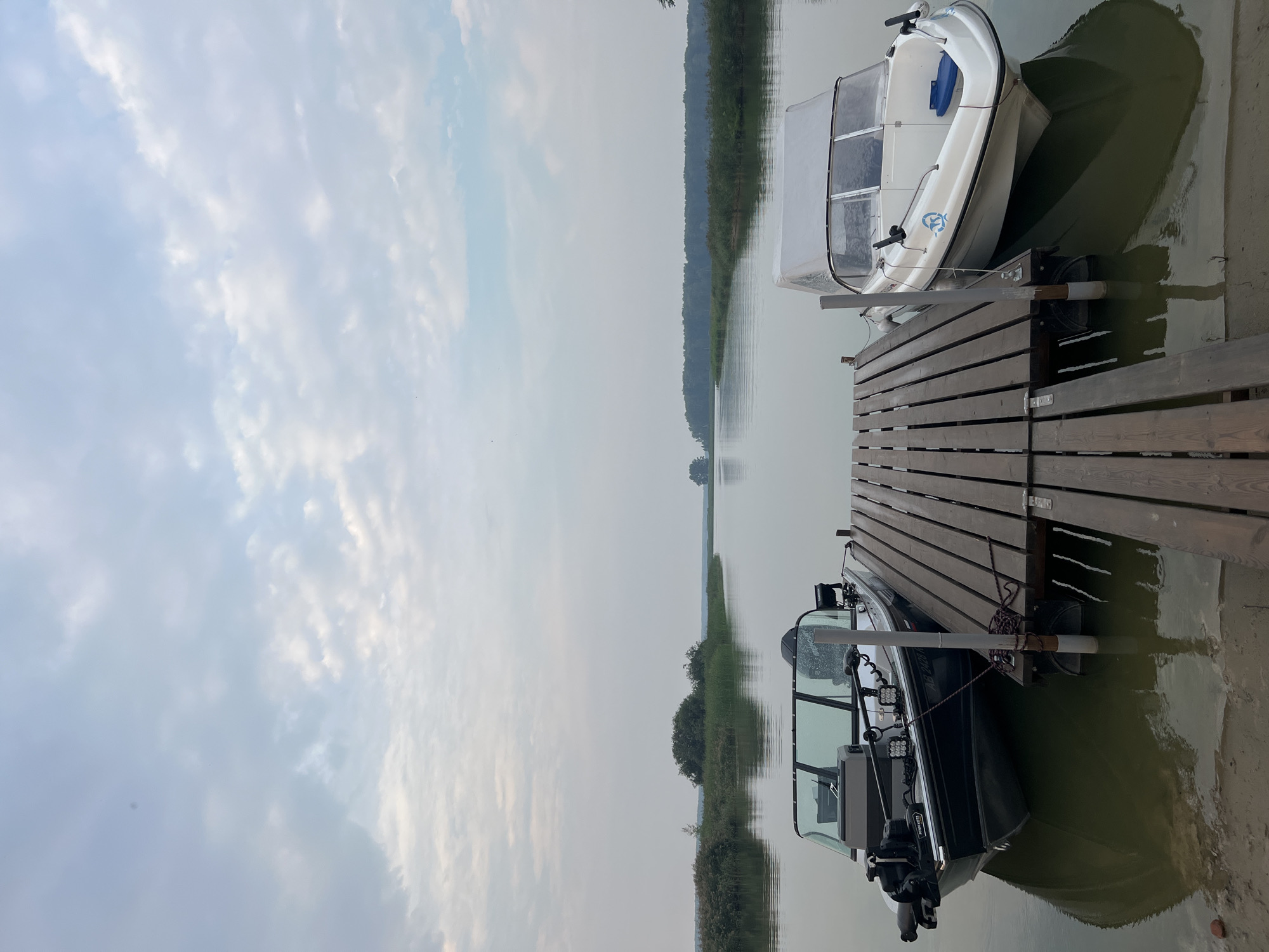 The image depicts a serene lake scene with two boats docked at a wooden pier. The sky is overcast with scattered clouds, casting a soft, diffused light on the landscape. The water is calm and reflects the surrounding greenery and the boats.
On the left side of the pier, a dark-colored fishing boat is moored. It has a windshield, a trolling motor at the bow, and what appears to be a Garmin fish finder mounted on the console. The boat's reflection is clearly visible in the water.
On the right side, a white boat with a canopy is also docked. It has a more streamlined design and a blue seat inside. Its reflection is also visible in the water.
The pier itself is made of weathered wooden planks and extends into the water. The surrounding environment features lush green reeds and trees along the shoreline, with a distant forested area visible in the background. The overall atmosphere is peaceful and tranquil.
The image is taken from a slightly elevated perspective, looking towards the water and the horizon. There are no people visible in the scene. The location is identified as Zav'yalovo, Russia, likely on a lake or a wide river.