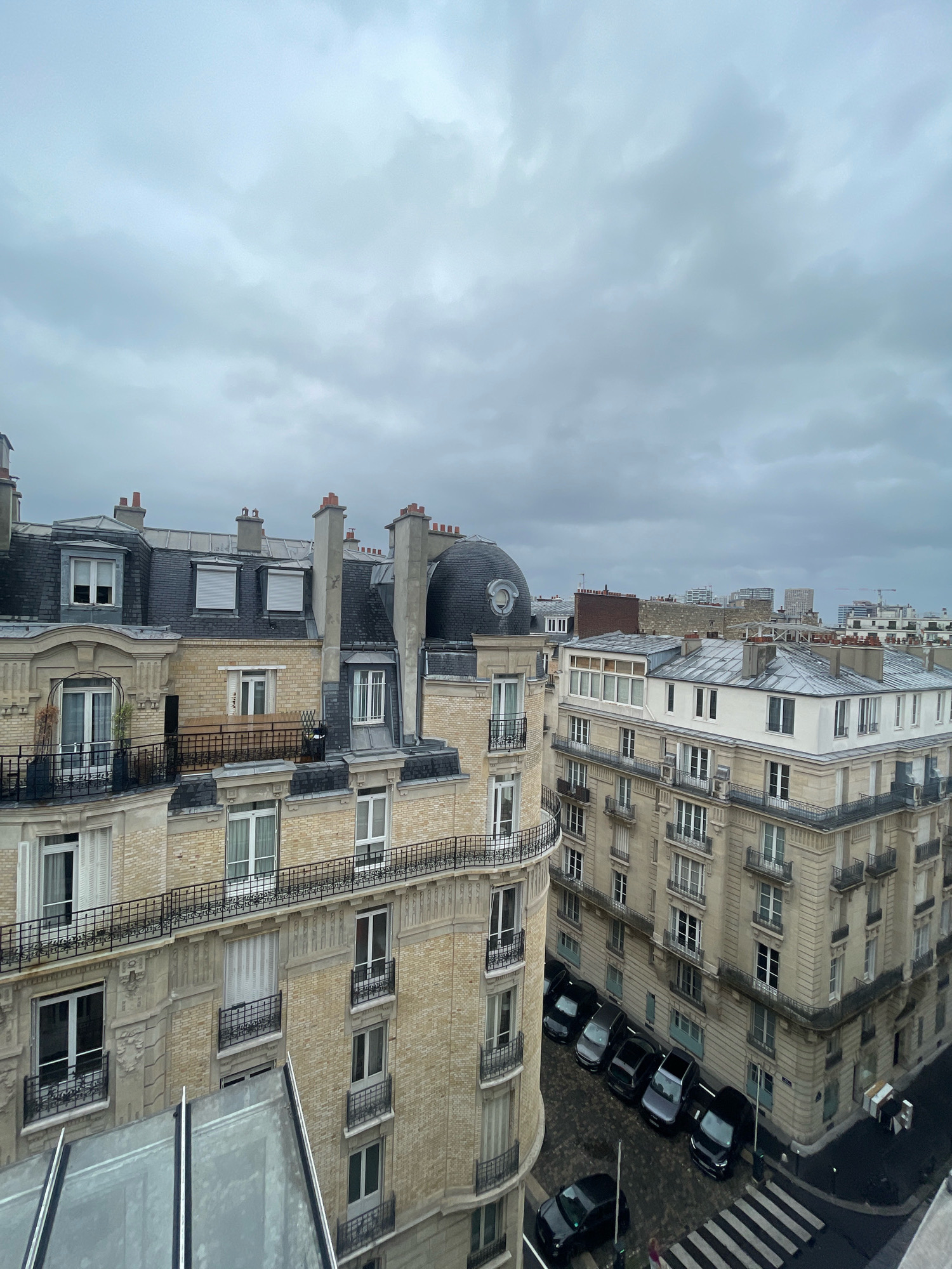 This image depicts a view of Parisian architecture on an overcast day. The buildings are characteristic of Haussmannian style, with light-colored stone facades, mansard roofs covered in slate, and numerous windows, many with decorative balconies and iron railings. Chimneys are visible on the rooftops. Several vehicles, including black sedans and SUVs, are parked along the street below, which features a pedestrian crosswalk. The sky is heavily clouded, suggesting a gray or rainy day. The scene appears to be a residential area within the city of Paris, France, based on the architectural style and urban setting.