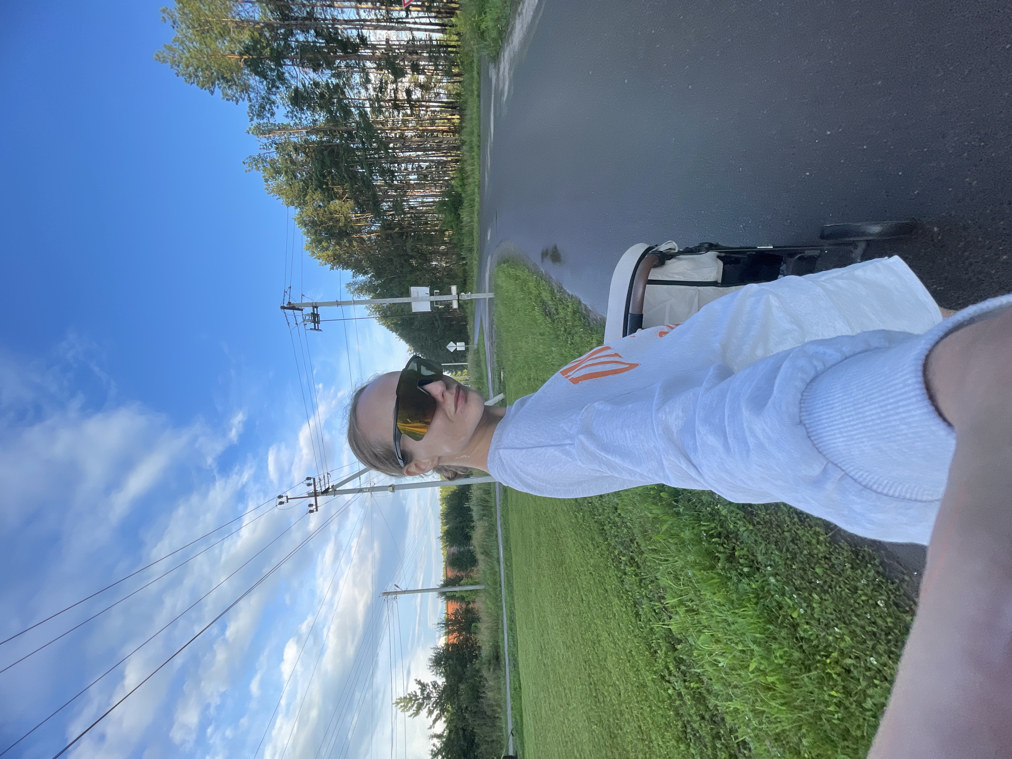 A woman with blonde hair pulled back into a bun, wearing large, dark sunglasses and a light-colored sweatshirt with orange lettering, is walking down a paved road in Podshivalovo, Russia. She is pushing a stroller beside her. The road is wet, with puddles visible. To the left of the road is a lush green field. To the right, a dense line of tall pine trees borders the road. In the background, there are power lines supported by utility poles, a few houses, and a bright blue sky with scattered white clouds. The scene appears to be during the late afternoon or early evening, given the lighting.