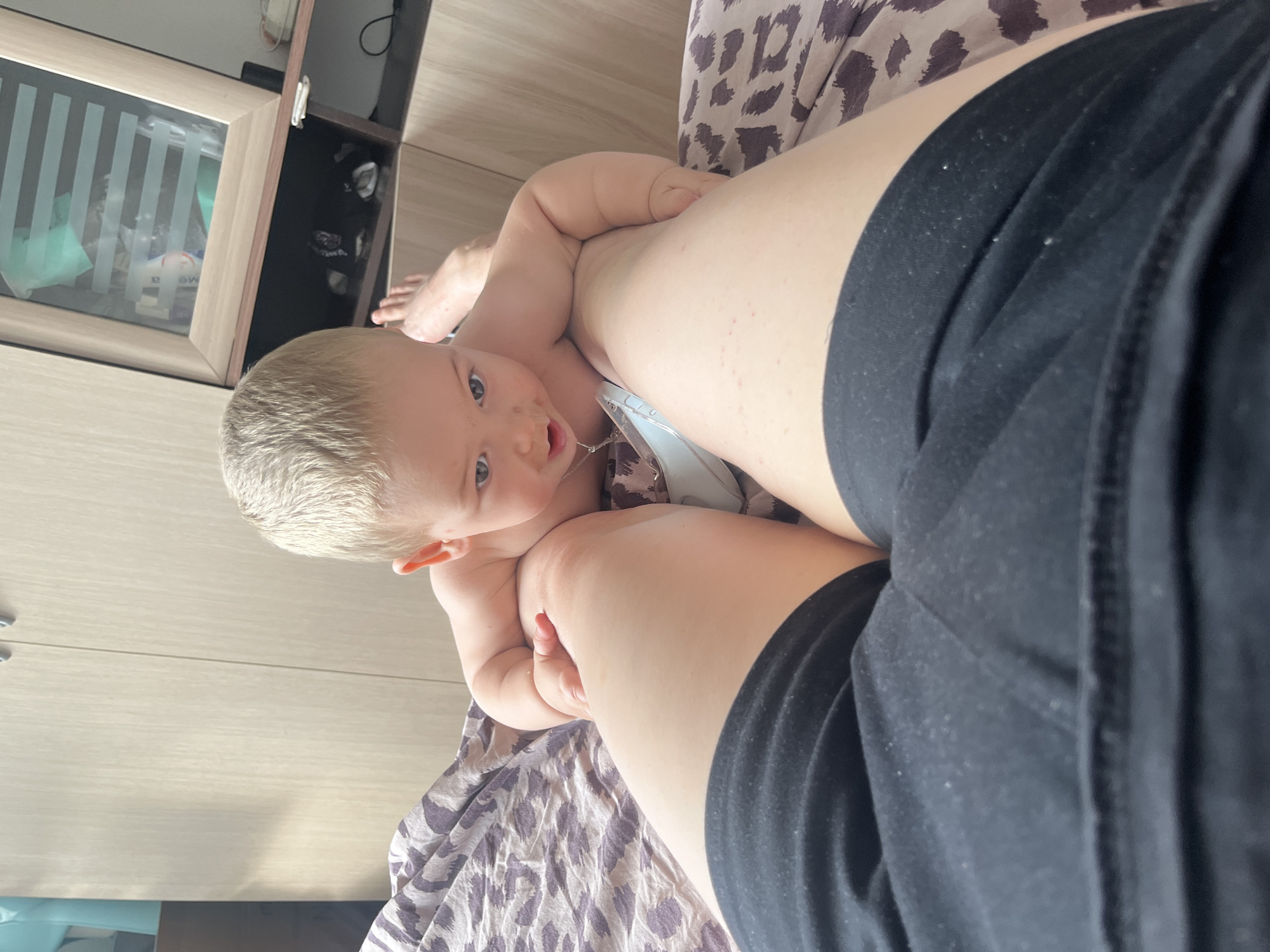 The image shows a baby's face, with blonde hair and blue eyes, looking up at the camera. The baby is positioned between the legs of an adult, whose black shorts are visible in the foreground. The adult's legs are also visible, with some redness on the skin. The baby is lying on a patterned fabric, possibly a blanket or bedding. In the background, there is a wooden cabinet with a glass door, revealing items inside. The scene appears to be indoors, likely in a home setting. The lighting suggests it could be daytime, but there are no clear indicators of the time of day or weather. The baby's expression is one of curiosity and engagement. The location context is Chelyabinsk, Russia. There is no visible text in the image.