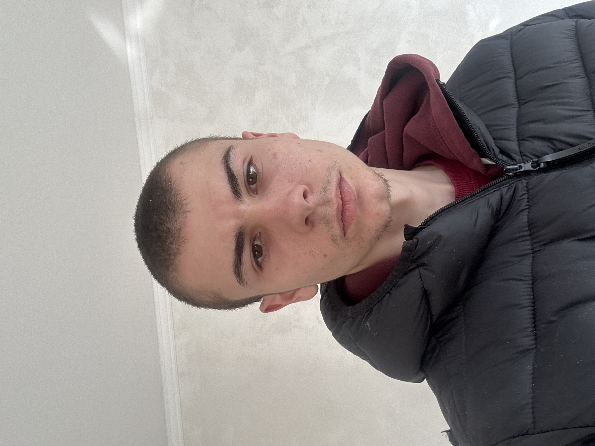 A close-up, angled portrait shot of a young man with a shaved head indoors. The man has brown eyes and light skin, with some facial hair growth. He is wearing a maroon hoodie beneath a black puffer jacket, partially zipped. The background is a plain, light-colored wall. The image appears to be taken in a room with neutral-toned walls. The location cannot be determined to be Plovdiv, Bulgaria, based solely on the given image.