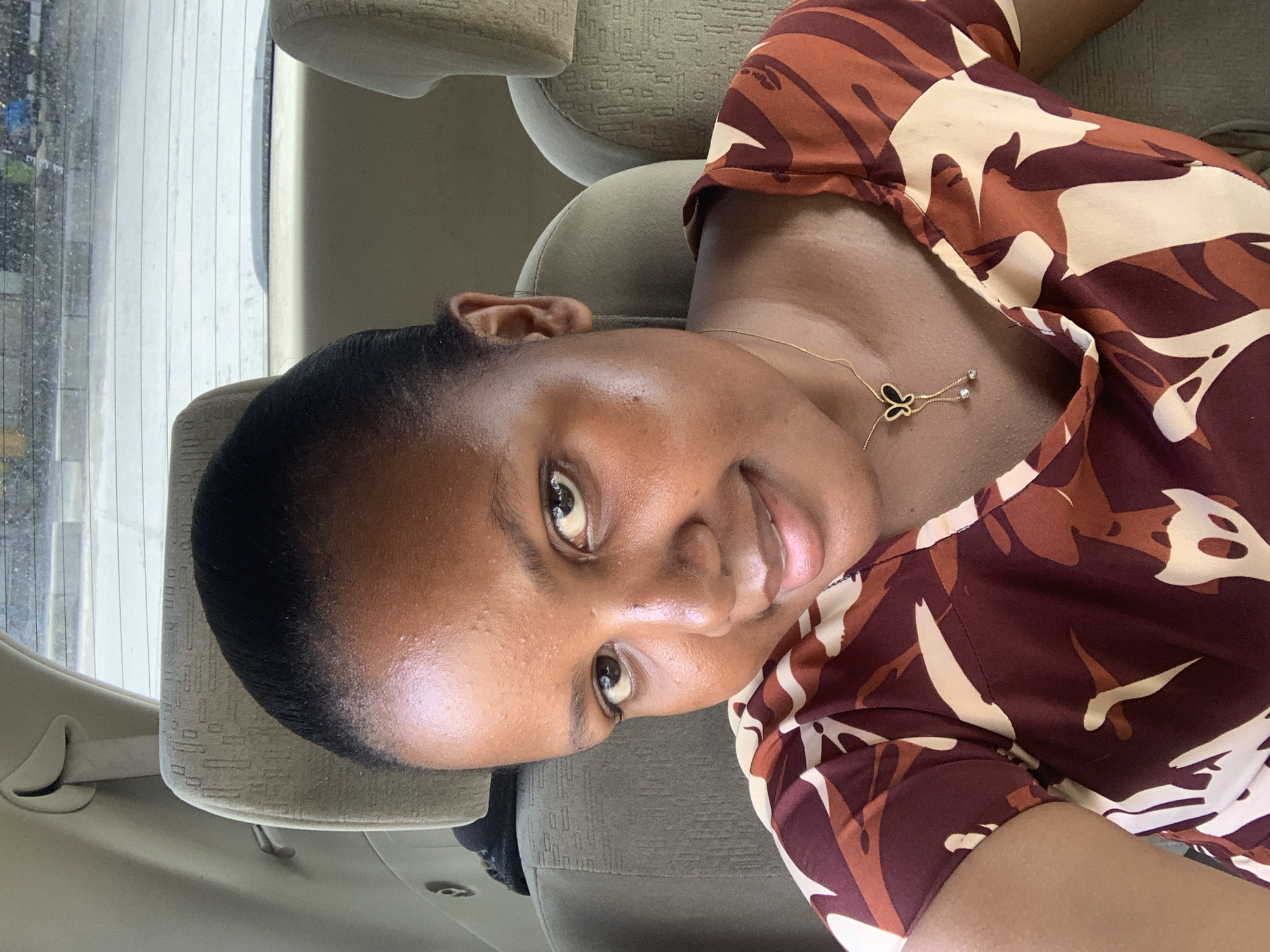 A dark-skinned woman with short black hair is sitting in a car. She is wearing a brown and beige patterned top and a gold necklace with a butterfly pendant. She is looking at the camera with a slight smile. The interior of the car is beige. The car window is visible in the background, with what looks like a street outside.
It is impossible to confirm the specific location in Dar es-Salaam, Tanzania from this image alone. Additional information such as landmarks visible outside the car window would be needed.