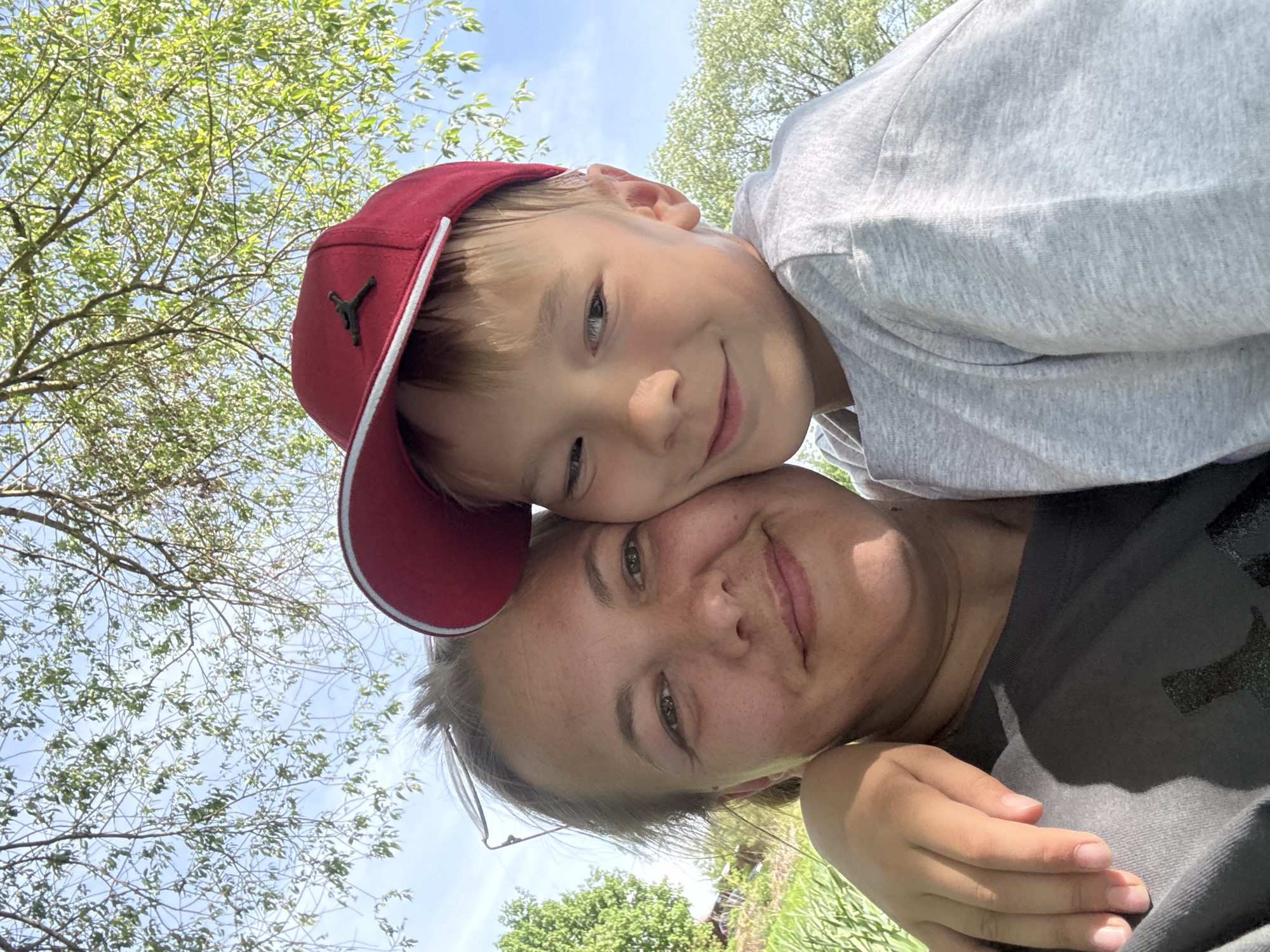 A selfie-style photograph captures a woman and a young boy outdoors, likely in the Синьково, Russia area, suggested by the presence of lush vegetation, trees with green leaves, and clear sky. The woman is in the foreground, with the boy leaning on her. The boy is wearing a red baseball cap with a black logo. Both are smiling, indicating a friendly and happy interaction. The background features branches of trees against a sky with little visible cloud cover.