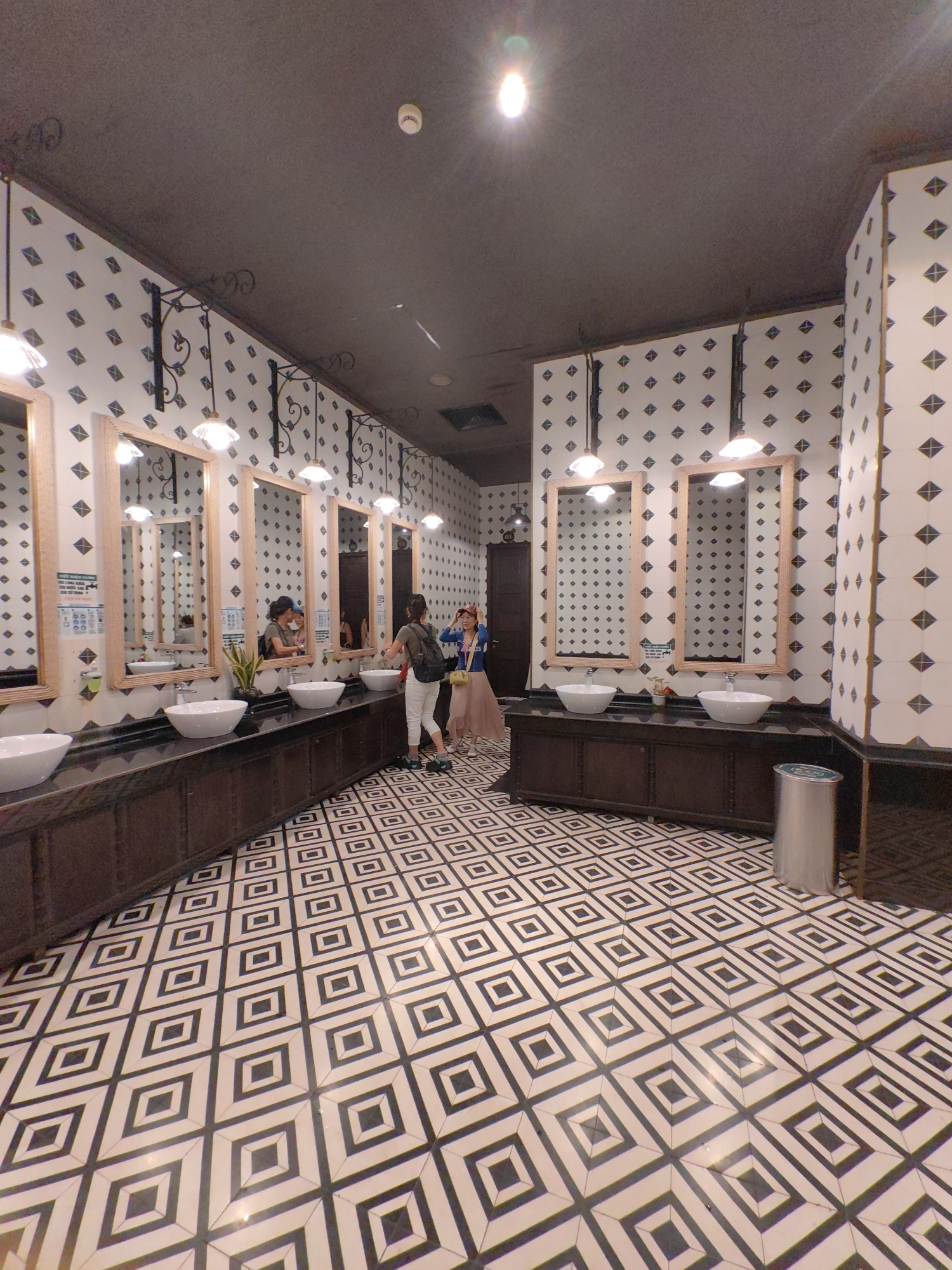 The photograph shows the interior of a restroom in Bai Chay Ward, Vietnam. The room features a black and white geometric tile floor and walls tiled with a pattern of small black diamonds on a white background. The ceiling is dark. There are multiple vanity stations, each with a light wood-framed mirror, a white vessel sink, and a dark-colored cabinet below. Several people are present: two women stand near the center of the room, and another woman is at one of the vanities. A metal trash can stands near the corner of the room.