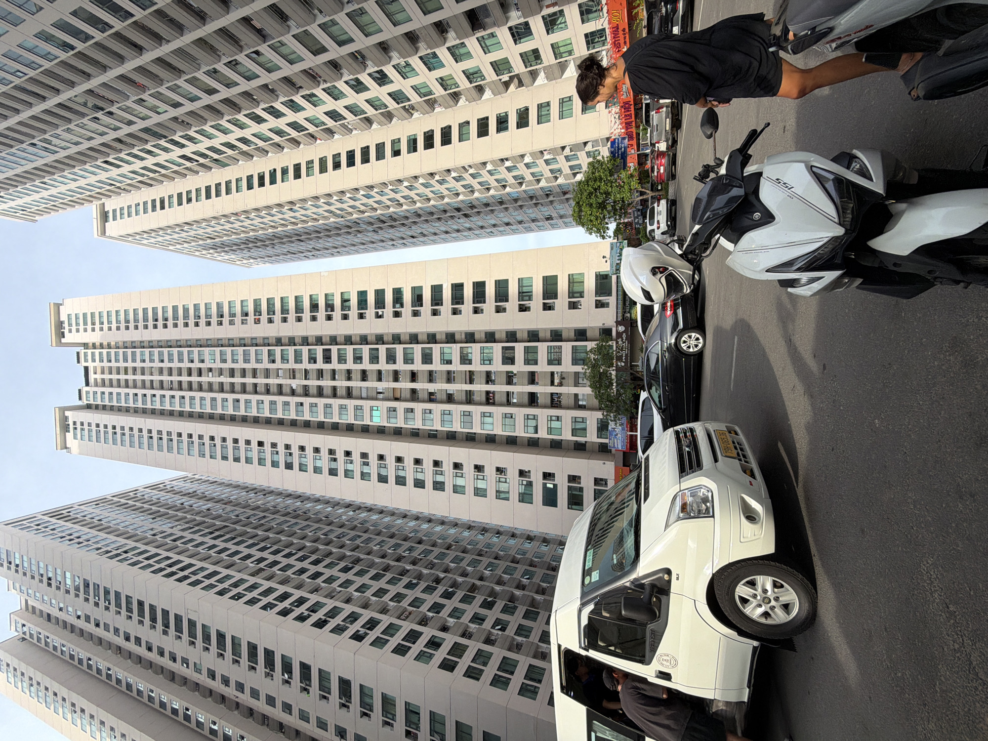 A wide shot captures a street scene in Bac Nha Trang Ward, Vietnam, dominated by towering apartment buildings. In the foreground, a white van is parked with its side door open, and a person wearing a black t-shirt and dark shorts is leaning into it. To the right of the van, a white motorcycle is parked, with a white helmet resting on its handlebars. A woman in a black t-shirt dress stands to the right of the motorcycle, facing away from the camera. Several other vehicles, including a black car and a silver SUV, are visible in the background, along with more of the apartment buildings. The sky is overcast.