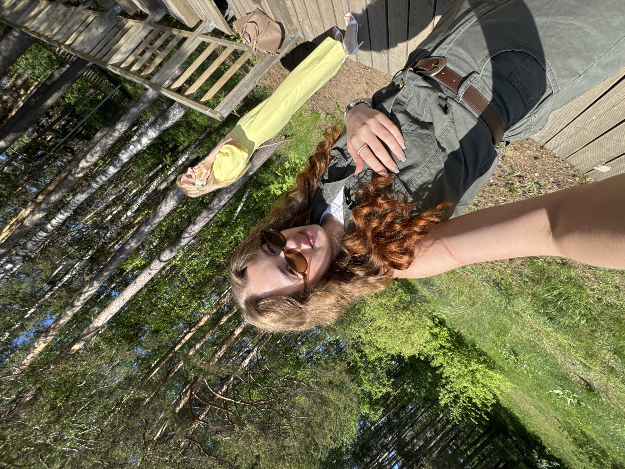 A selfie photograph shows two women outside on a sunny day, likely in the city of Chaikovsky, Russia. The camera angle is tilted to the left, with the ground and trees in the background.
One woman is close to the camera, partially in frame, wearing sunglasses, a green jacket, and possibly cargo pants with a brown belt. She has light brown hair with red curls. Her right hand is visible, displaying a ring on her ring finger.
The second woman is further away, fully visible, lying on a wooden surface wearing a yellow outfit. She has blonde hair and white high-heeled shoes. Next to her is a light brown purse.
The background consists of trees and greenery, with glimpses of the sky. A wooden structure resembling a staircase or railing is visible in the upper left. The foreground is a wooden deck. The scene conveys a casual, outdoor setting, possibly a park or recreational area.