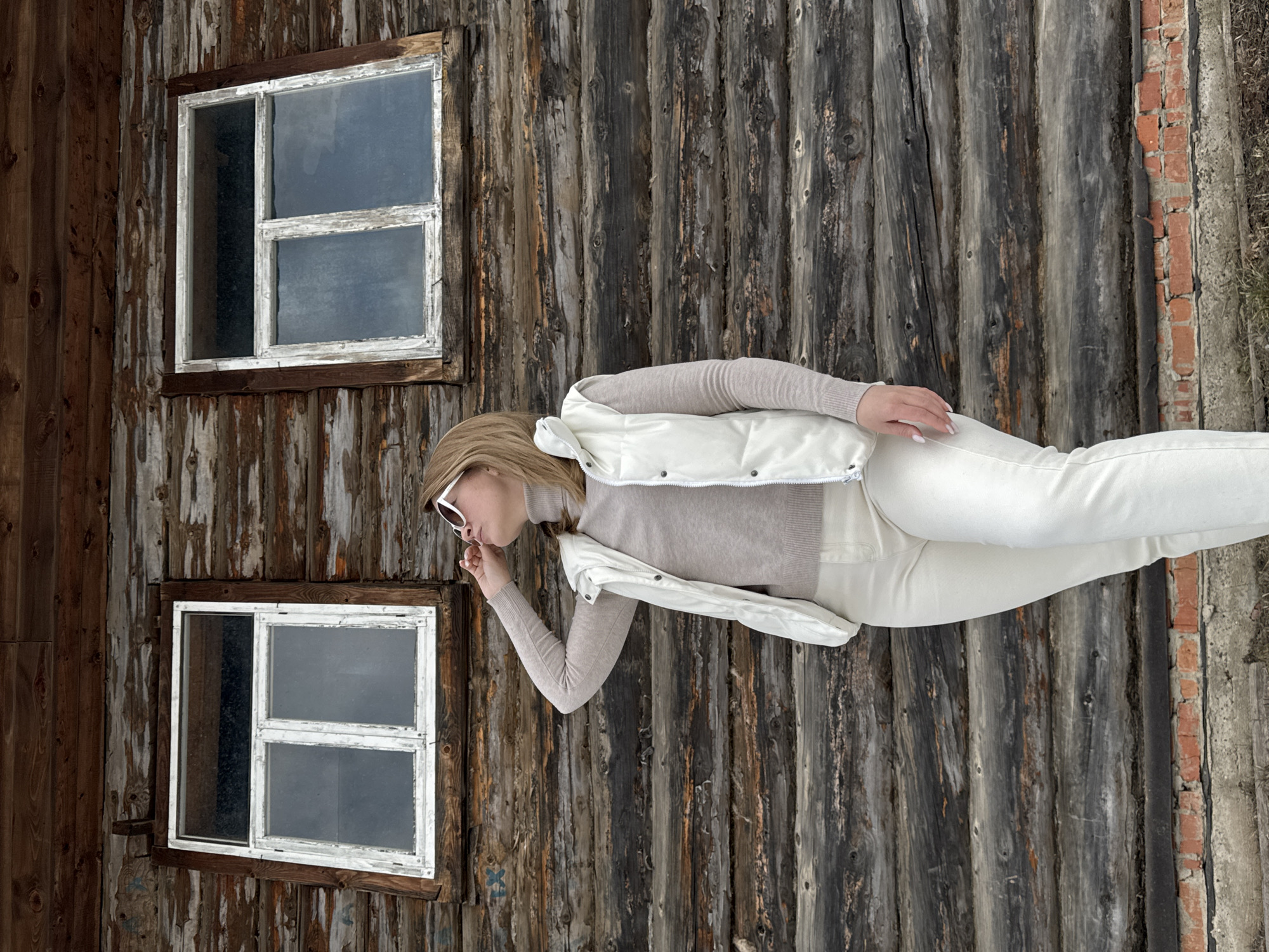The image depicts a woman standing in front of a wooden building in what appears to be a rural area. The woman is wearing a light beige sweater, white pants, and a white vest. She is wearing sunglasses and has blonde hair. She is positioned on the right side of the frame. The building behind her is constructed of dark, weathered wooden planks, featuring two windows with white frames. Adjacent to the wood structure is a brick area. It is likely that the location is Amaneevo, Russia as was specified.