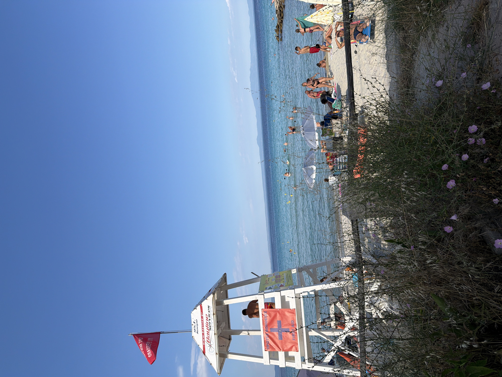 This media file depicts a sunny day at a crowded beach in Kassandra, Greece. The scene is dominated by a clear, bright blue sky and the calm, blue waters of the sea. The beach itself is sandy, with a line of beach umbrellas and lounge chairs occupied by people sunbathing and swimming. In the foreground, a white lifeguard tower stands tall, with a person visible inside. A red flag with white lettering flies from a pole near the tower. Several signs are attached to the lifeguard tower, including one with "Bayline Services" written on it and another displaying a red cross symbol with the words "FIRST AID". The presence of numerous people enjoying the beach suggests a popular tourist destination during peak season. The overall mood is relaxed and summery, with the bright sunlight and blue water contributing to a sense of leisure and vacation. The vegetation in the foreground, with its dry grasses and small pink flowers, adds a natural element to the scene. The time of day appears to be midday, given the bright sunlight and long shadows.