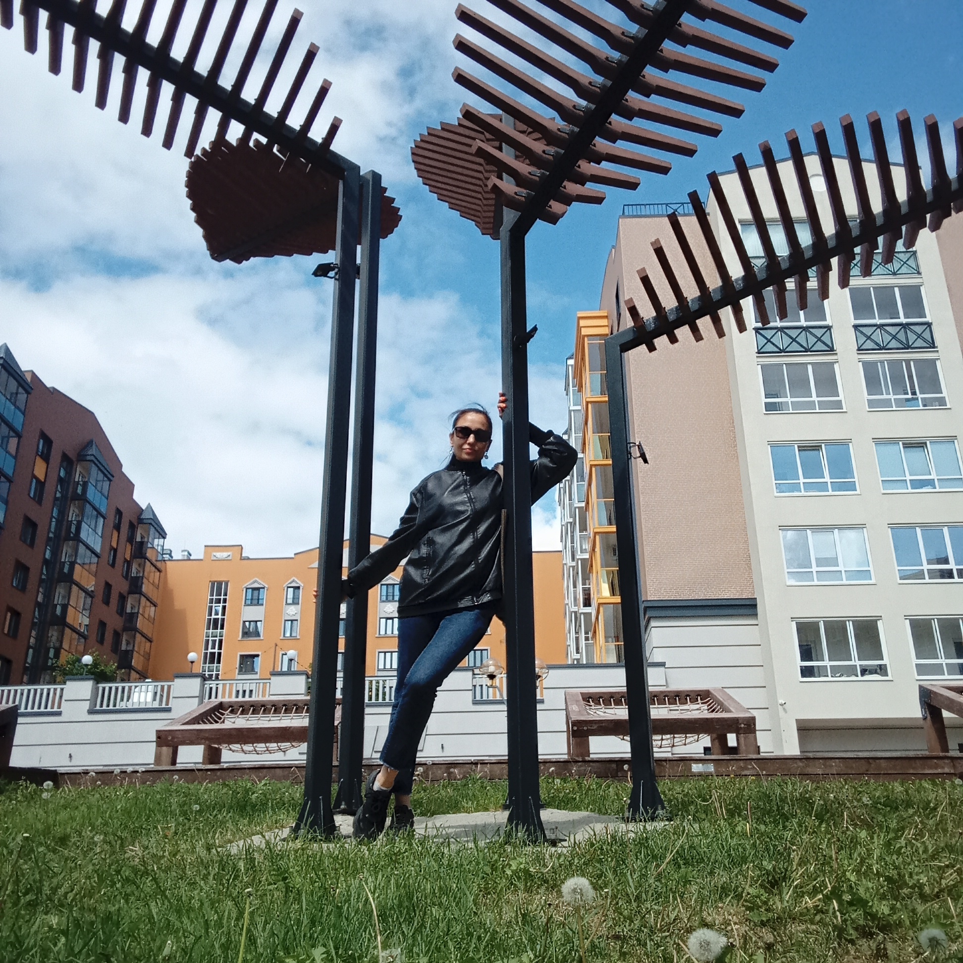 A wide-angle, low-angle shot captures a woman posing casually in an outdoor urban setting, likely a residential courtyard in Pervomaysky, Russia. The woman is centrally framed, leaning against a dark metal post of a modern outdoor structure, possibly a pergola or decorative arbor, which features horizontal slatted wooden beams overhead. She is looking directly towards the camera with a confident and relaxed expression, wearing dark sunglasses.
She is dressed in a black leather jacket, blue jeans, and black athletic shoes. Her arms are positioned on the structure, with her right arm bent and resting on a beam, and her left hand gripping a vertical post. Her left leg is slightly bent and crossed in front of her right.
The foreground consists of green grass dotted with white dandelions. In the background, several multi-story apartment buildings with contemporary architecture are visible under a bright, partly cloudy blue sky. The buildings display varied facades in shades of light beige, orange-brown, and brick-red, featuring numerous windows and some glass-enclosed balconies. The overall scene suggests a well-maintained, modern residential area. The clear sky and distinct lighting indicate it is daytime, likely morning or early afternoon. No visible text is present in the image.