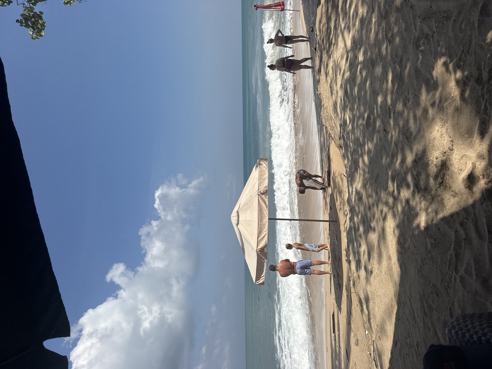 This is a horizontal, eye-level, medium shot of a beach in Kuta, Indonesia, on a sunny day with a clear blue sky and scattered white clouds. The foreground is dominated by sandy beach with dappled shadows cast by unseen trees. In the mid-ground, a large, light-colored beach umbrella stands center-left.
To the left of the umbrella, a man and a boy stand facing the ocean. The man is shirtless, wearing blue patterned shorts, and the boy is also shirtless and wearing blue patterned shorts. They are standing close to the water's edge.
To the right of the umbrella, another man is bending over on the sand. Further to the right, two women in bikinis are standing and facing the ocean. One woman wears a dark bikini and the other wears a black bikini.
The ocean is a turquoise color with white-capped waves breaking on the shore. In the far right of the image, a red and white beach umbrella is visible. The overall scene depicts a typical beach day in a tropical location.