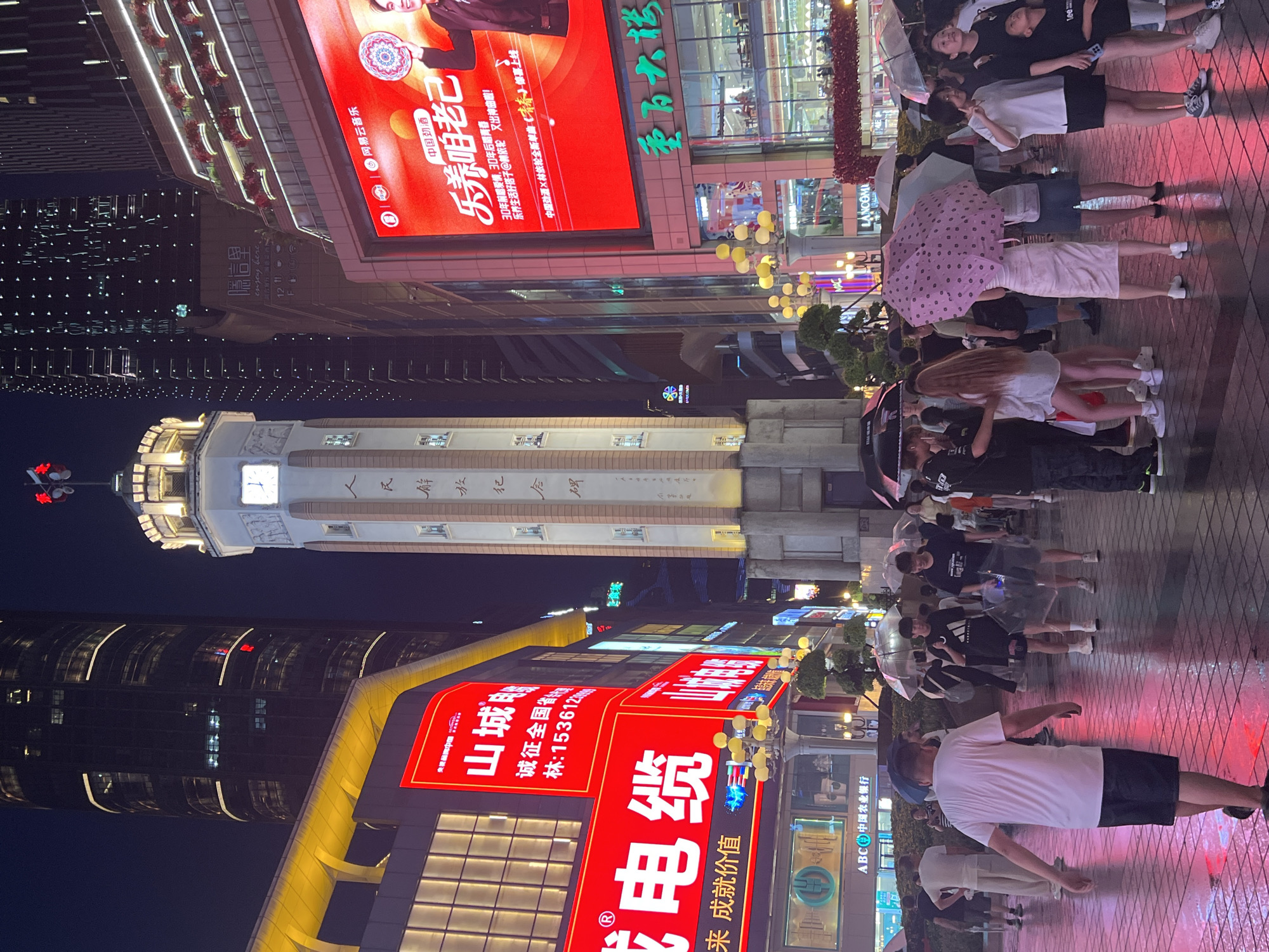 In Yuzhong District, China, at night, a city street scene is captured with illuminated buildings and a crowd of people walking under umbrellas on a wet pavement. A prominent clock tower stands in the background, adorned with vertical Chinese inscriptions. To the left, a large red advertisement displays Chinese characters and a phone number. To the right, a massive red digital billboard features an image of a man holding a decorative plate and Chinese text. The street is lined with modern buildings, some with illuminated windows and balconies adorned with red decorations. People are dressed in casual attire, with many holding umbrellas, suggesting it is raining or has recently rained, as evidenced by the wet, reflective pavement. The atmosphere is bustling and brightly lit by the surrounding advertisements and building lights.
